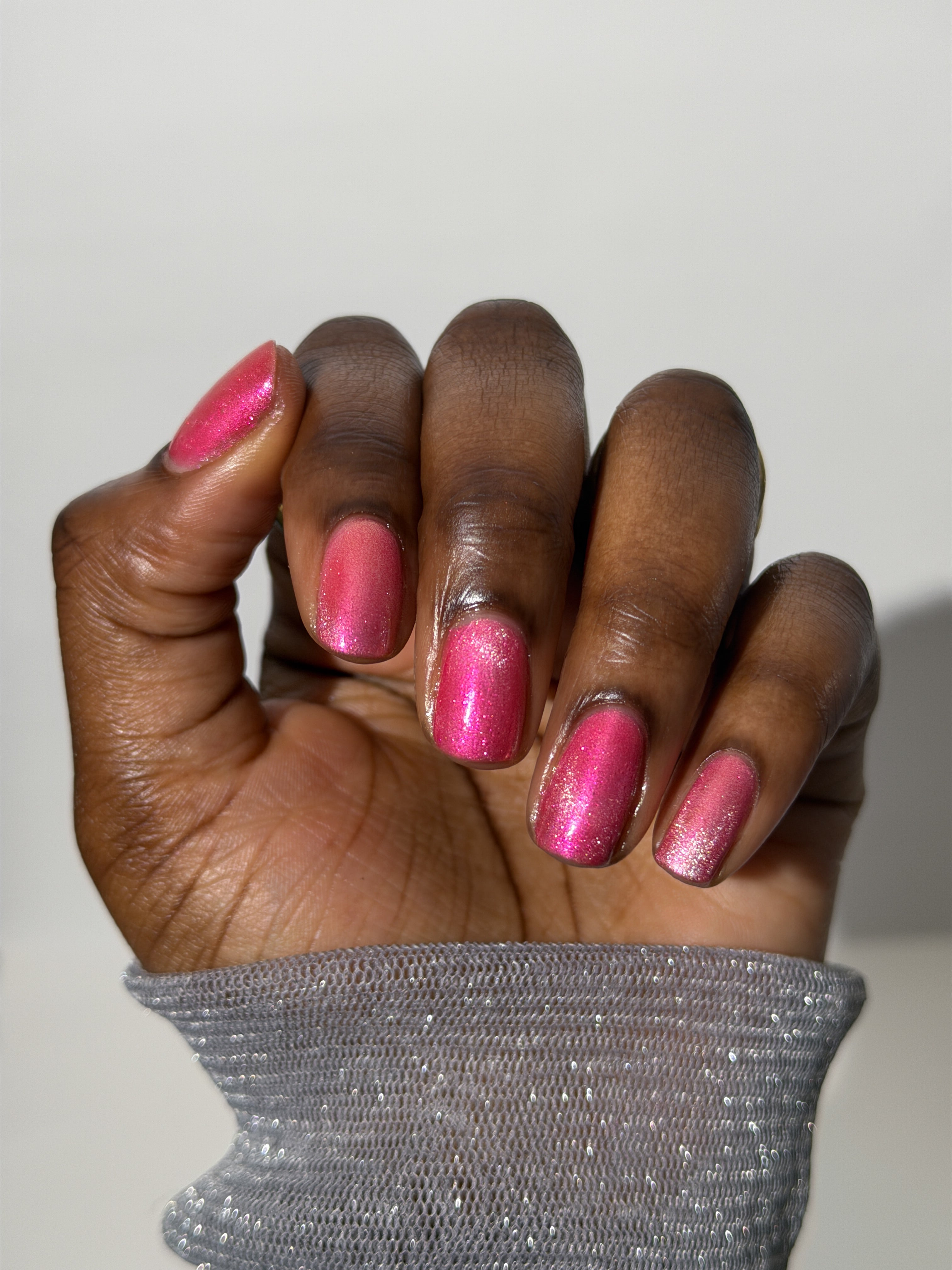 Lovey Dovey by Glitch Lacquer on five round nails showing a subtle magnetic stripe, warm pink polish with pink-to-gold reflective shimmer, deep skin tone, studio lighting.