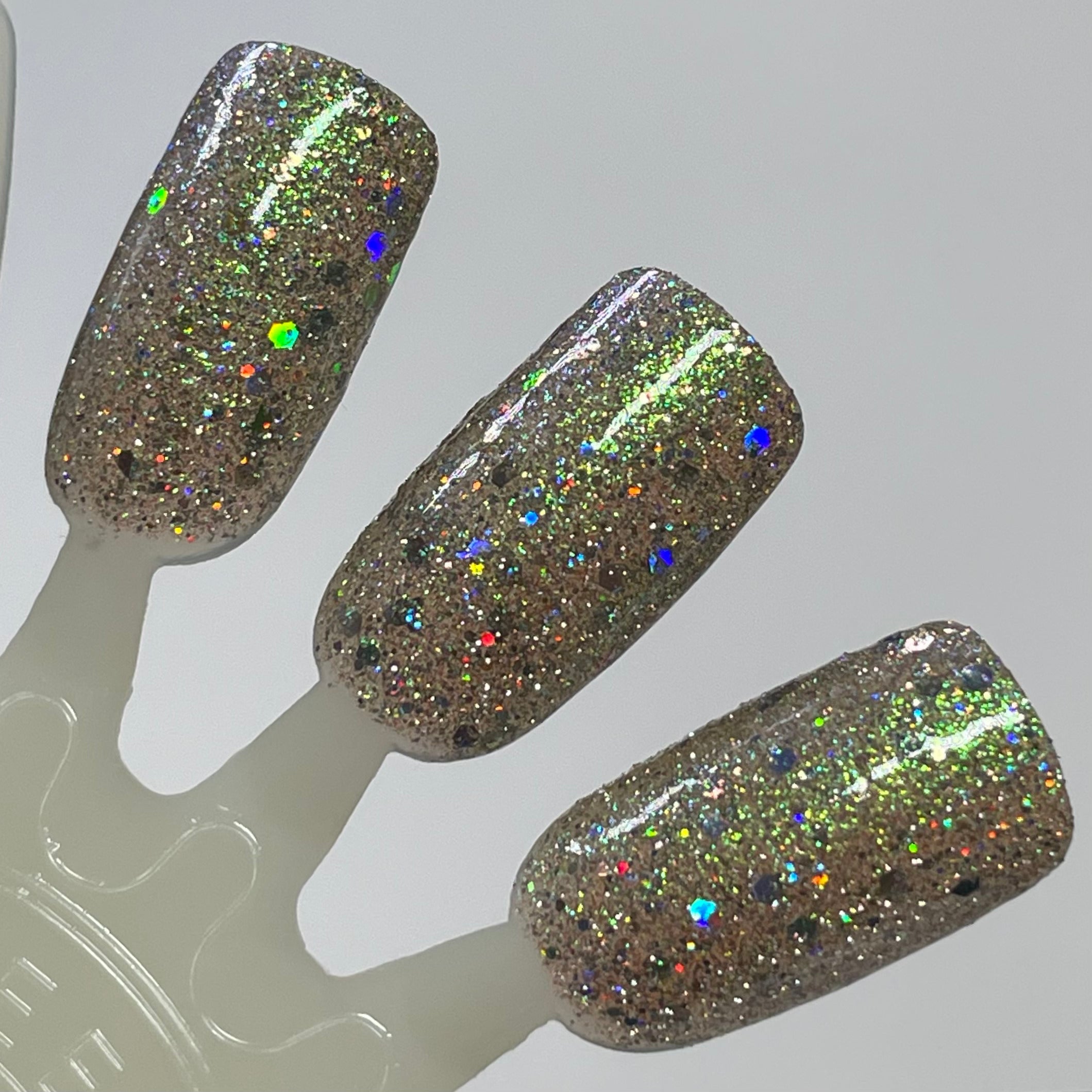 Thyme to Shine by Dam Nail Polish, warm taupe-greige base with green-to-gold shimmer shift, mixed fine micro-glitter and multicolour hex-cut holographic flakes under direct flash, swatch stick