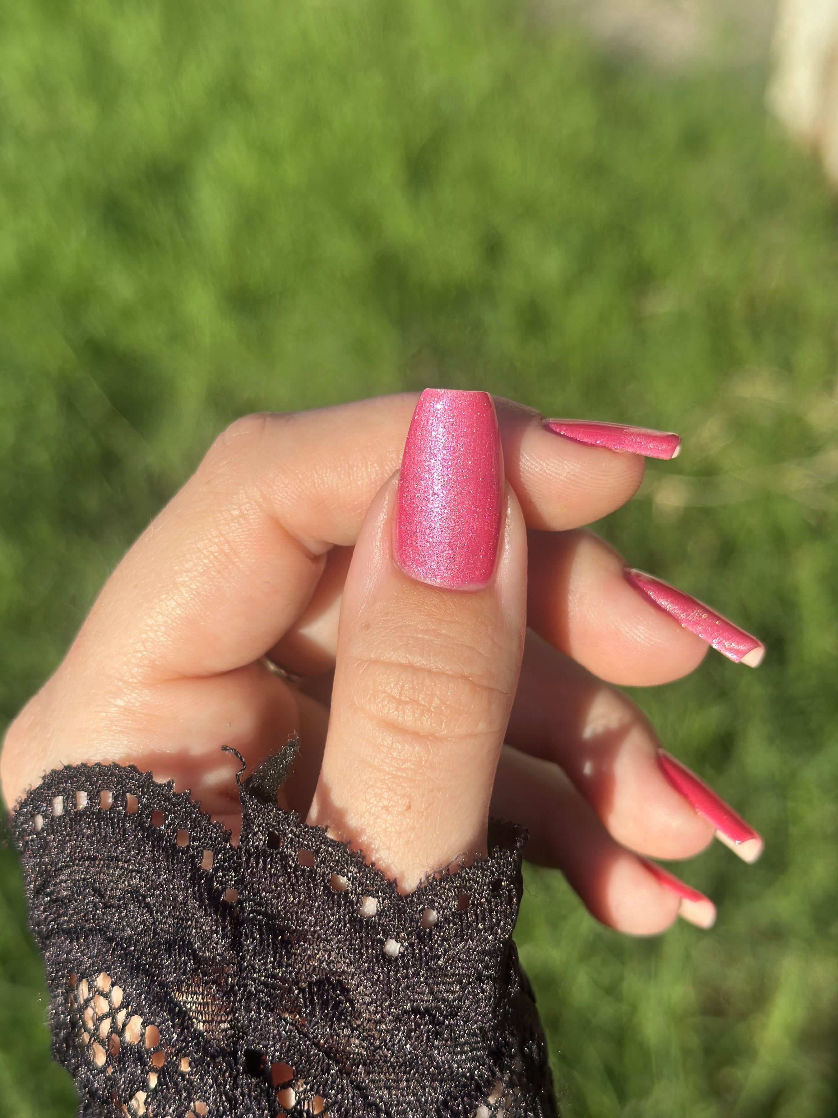 Lovey Dovey by Glitch Lacquer on five rectangular nails worn with a black lace cuff, warm pink polish with pink-to-gold reflective shimmer, light-medium skin tone, bright daylight.