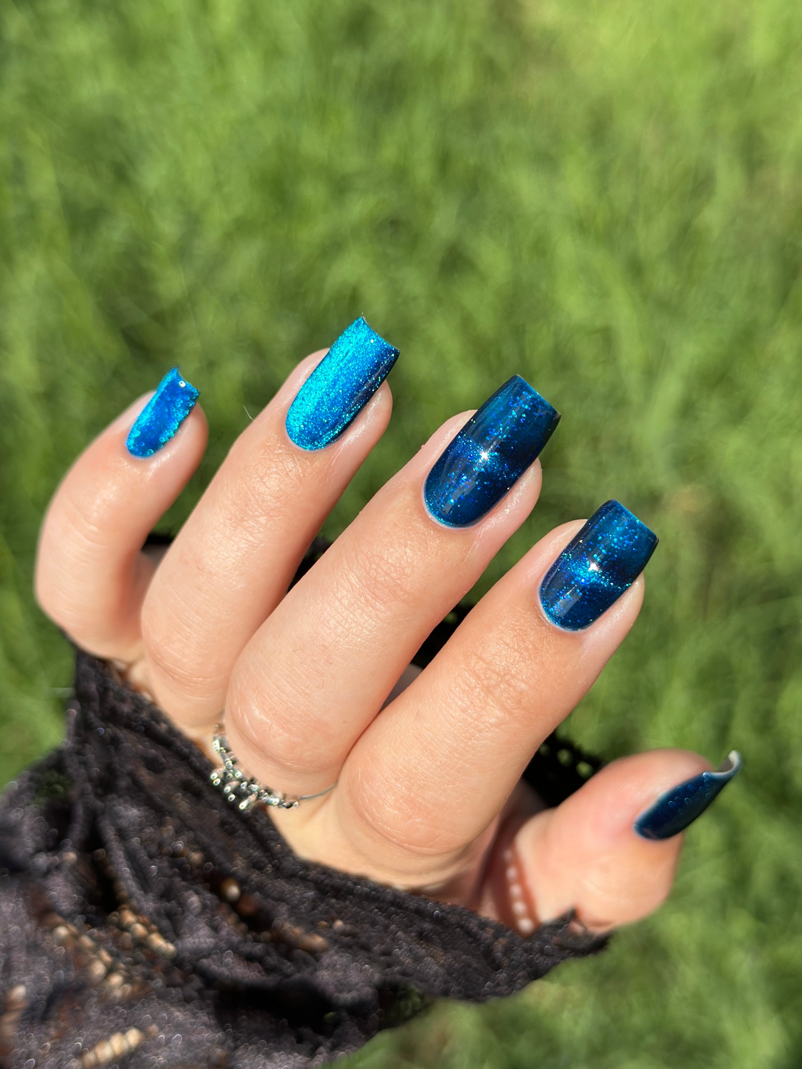 Heart of the Ocean by Glitch Lacquer on five square nails showing magnetic stripes across the middle and ring fingers, deep blue jelly with blue glitters, light-medium skin tone, bright daylight.