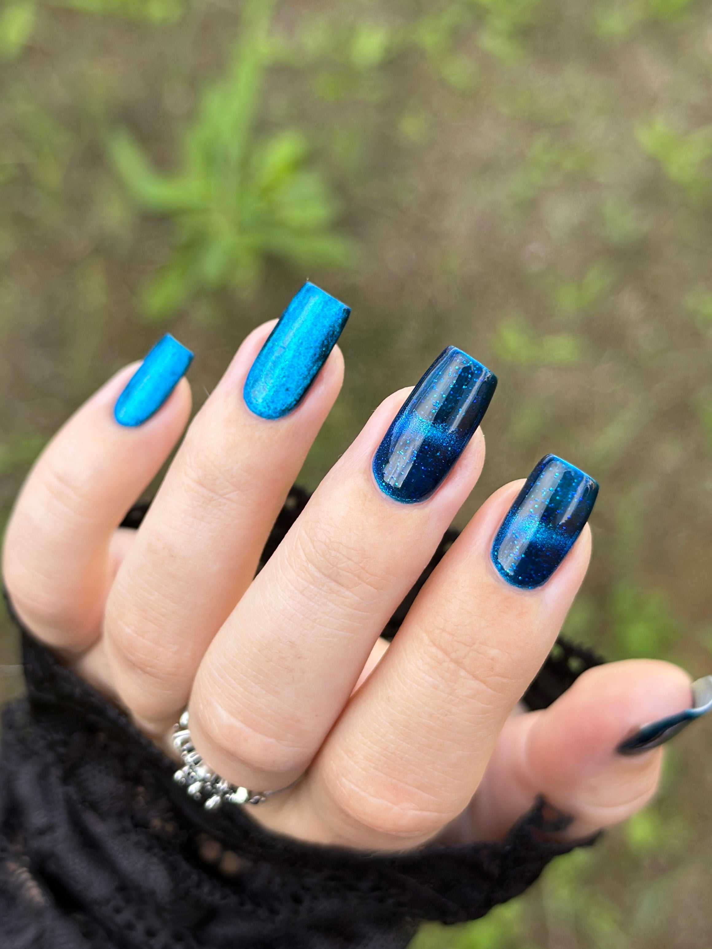 Heart of the Ocean by Glitch Lacquer on five square nails comparing magnetized and unmagnetized finishes, deep blue jelly with blue-to-purple magnetic shift and blue glitters, light-medium skin tone, bright daylight.