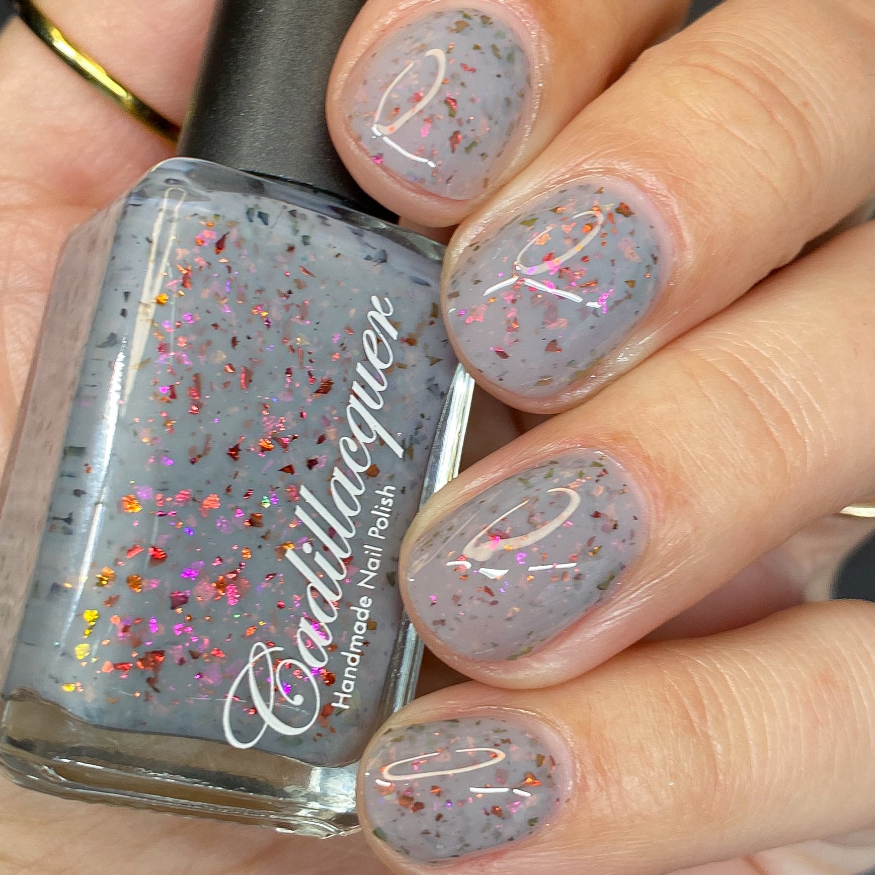 Cadillacquer Daydreaming grey red chameleon iridescent crelly nail polish, bottle held with four-nail swatch, light skin, bright daylight lighting