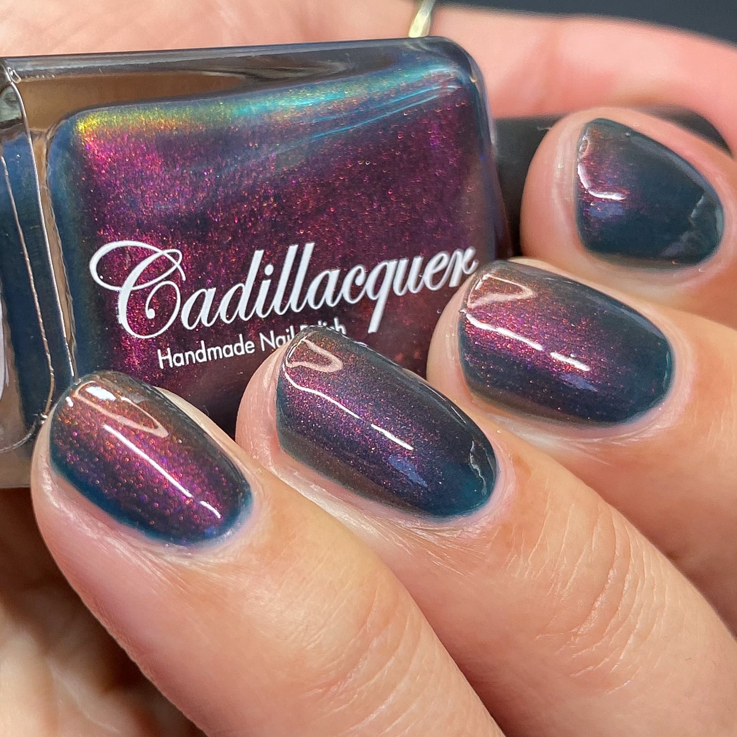Multichrome shimmer Cadillacquer nail polish in Fear What You Will Become, shown in bottle held in hand with four swatched nails, base shifts from murky dark teal to magenta and copper-orange with angle, light skin tone, studio lighting, swatched by @heeymercedes