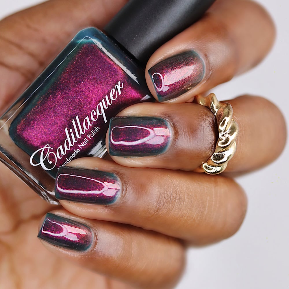 Multichrome shimmer Cadillacquer nail polish in Fear What You Will Become, shown in bottle held against deep-toned hand with four swatched square nails displaying a dark murky teal base shifting to deep magenta and bright pink-magenta at angle, with a decorative gold twisted ring, under studio lighting, swatched by @lacquer_is_life