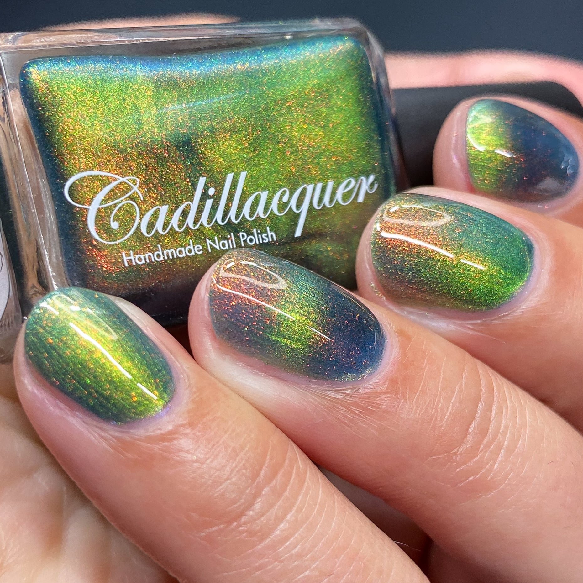 Magnetic multichrome Cadillacquer nail polish in I Found A Place, showing a bottle held in hand alongside four swatched short oval nails, with a deep teal base shifting to lime-green magnetic band and red-to-orange aurora shimmer, on light skin tone, under studio lighting, swatched by @heeymercedes