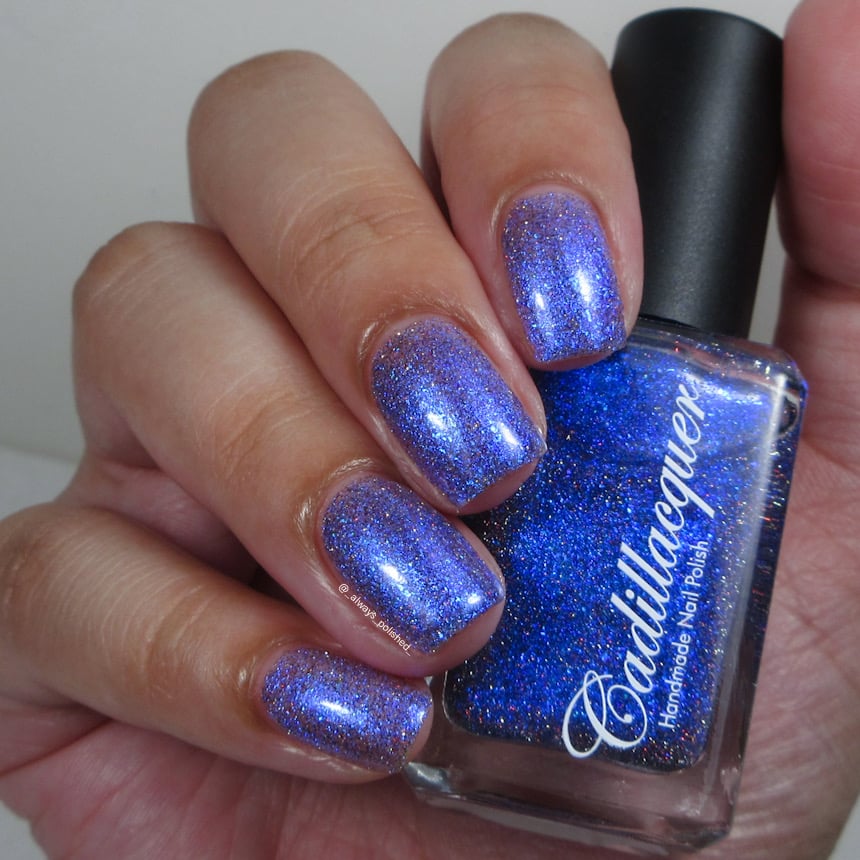 Shimmer, flakies, and holographic glitter Cadillacquer nail polish in I'll Be Right Back, shown on five squoval nails held alongside the bottle, nails read periwinkle-violet with scattered blue shimmer and tiny red and charcoal holographic sparks while the bottle face reads vivid royal cobalt blue, light skin tone, indoor warm lighting, swatched by @_always_polished_