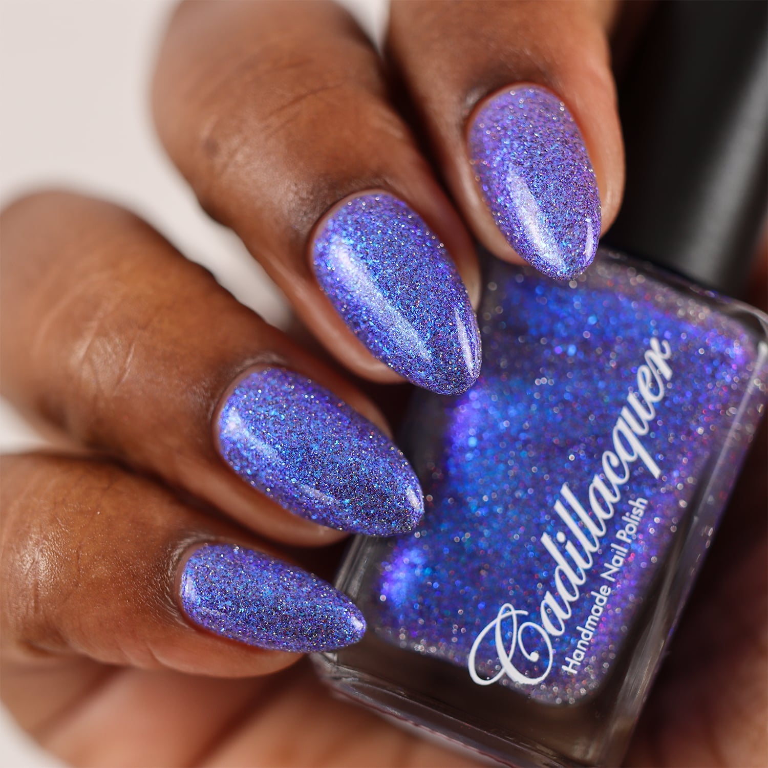 Shimmer, glitter, and micro-flake Cadillacquer nail polish in I'll Be Right Back, shown on four almond-shaped nails on rich skin tone alongside the bottle held in hand, the polish reads periwinkle-violet with dense blue micro flakes, electric blue shimmer flash, and scattered silver-charcoal and red holographic glitter sparks, studio lighting, swatched by @doseoflolade