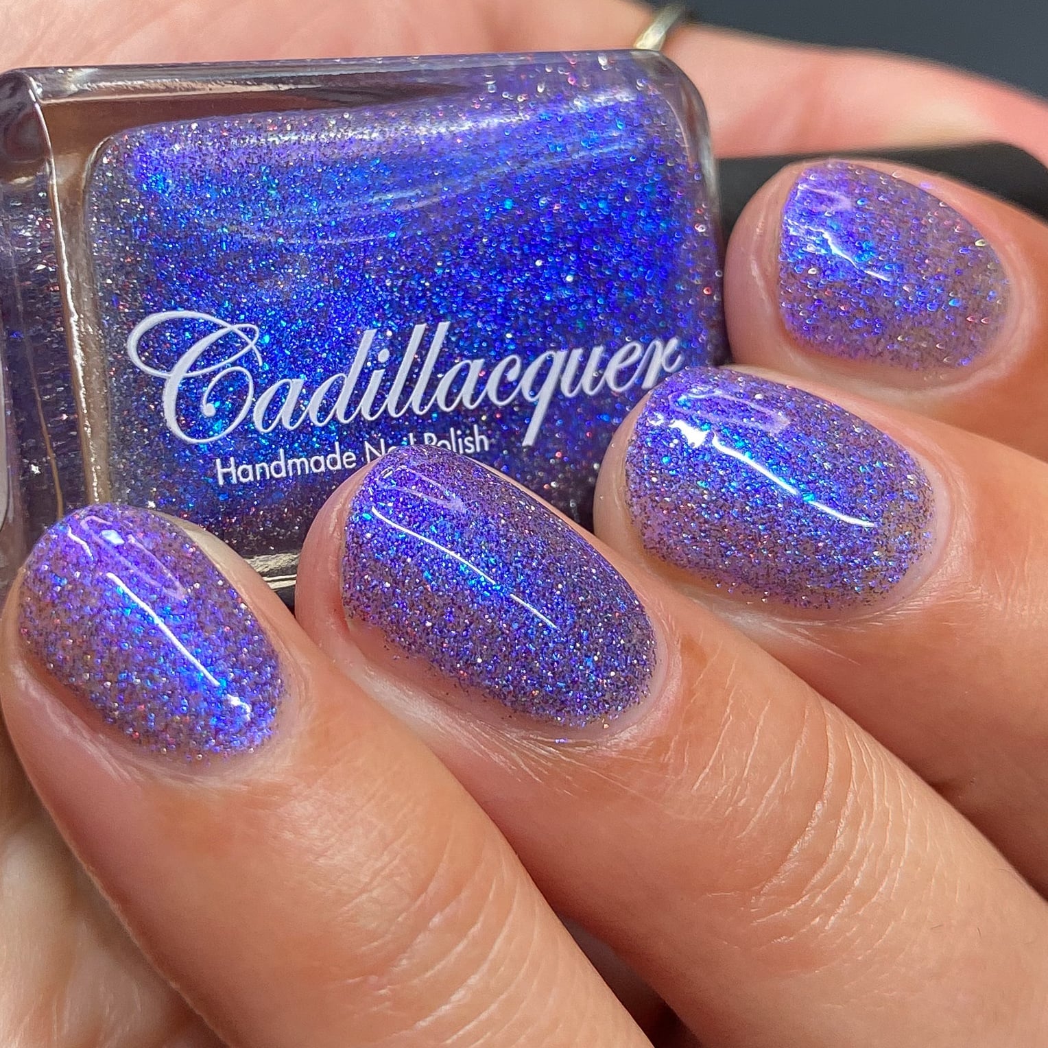 Holographic shimmer and micro-flake Cadillacquer nail polish in I'll Be Right Back, shown as bottle-in-hand with four swatched oval nails, polish is a dense blue-violet packed with blue micro flakes, royal blue shimmer flash, and scattered red and charcoal holographic glitter, light-medium skin tone, studio lighting, swatched by @heeymercedes