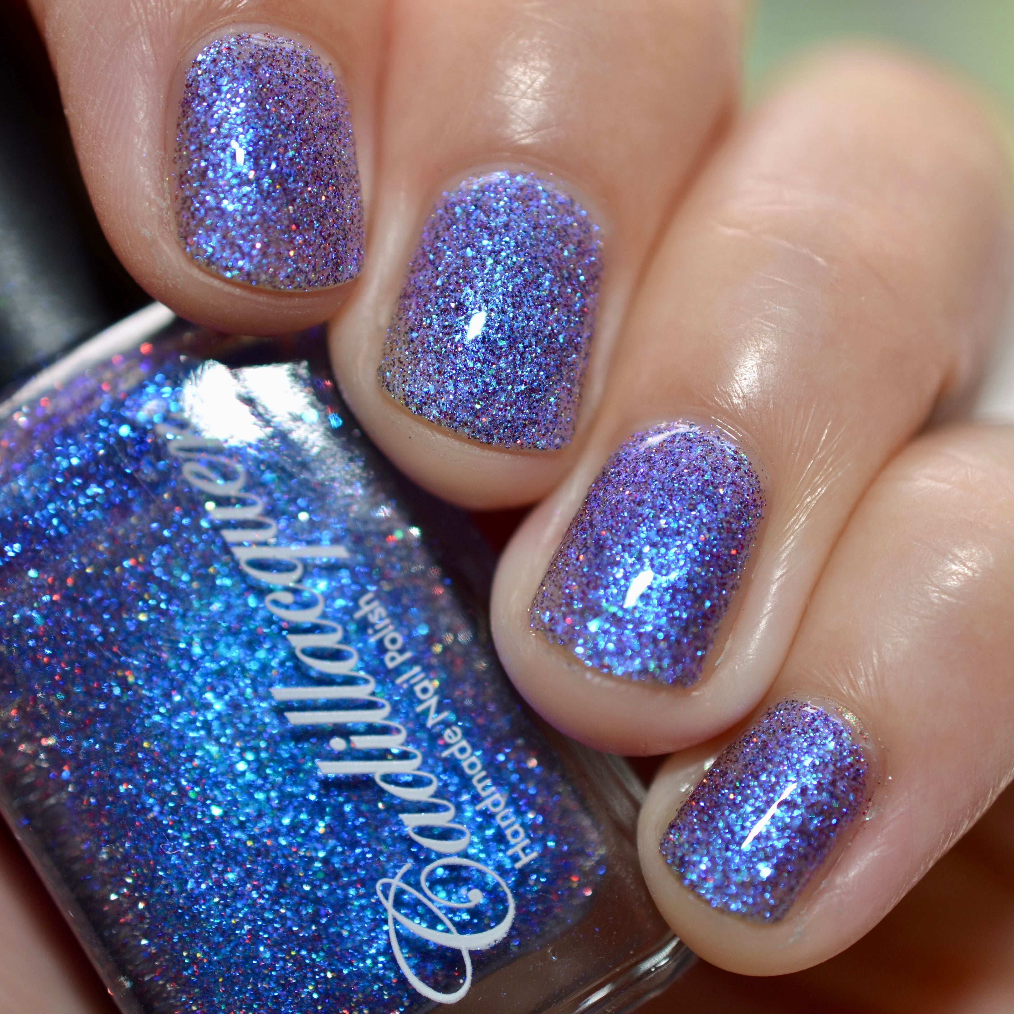 Shimmer, fine flake and reflective glitter Cadillacquer nail polish in I'll Be Right Back, shown as a bottle-in-hand swatch on four short natural nails, the clear base is packed with blue micro flakes and blue shimmer producing a periwinkle-violet tone on the nails, with red and charcoal reflective glitter sparks visible, the bottle face reads as vivid cobalt blue, light skin tone, studio lighting, swatched by @caitlinswatches