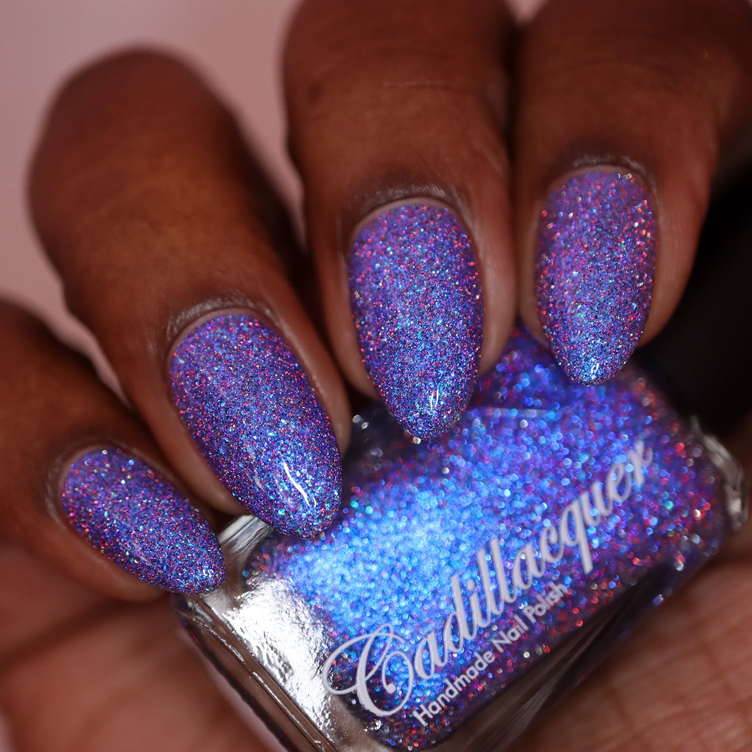 Shimmer, micro-flake and reflective glitter Cadillacquer nail polish in I'll Be Right Back, swatched on five oval nails held alongside the bottle, nails show a blue-violet sheer base packed with bright blue micro flakes, sky-blue shimmer, and a dense scatter of red-pink and charcoal holographic micro glitter, bottle face reads vivid cornflower blue under direct studio light, rich skin tone, studio lighting, swatched by @doseoflolade