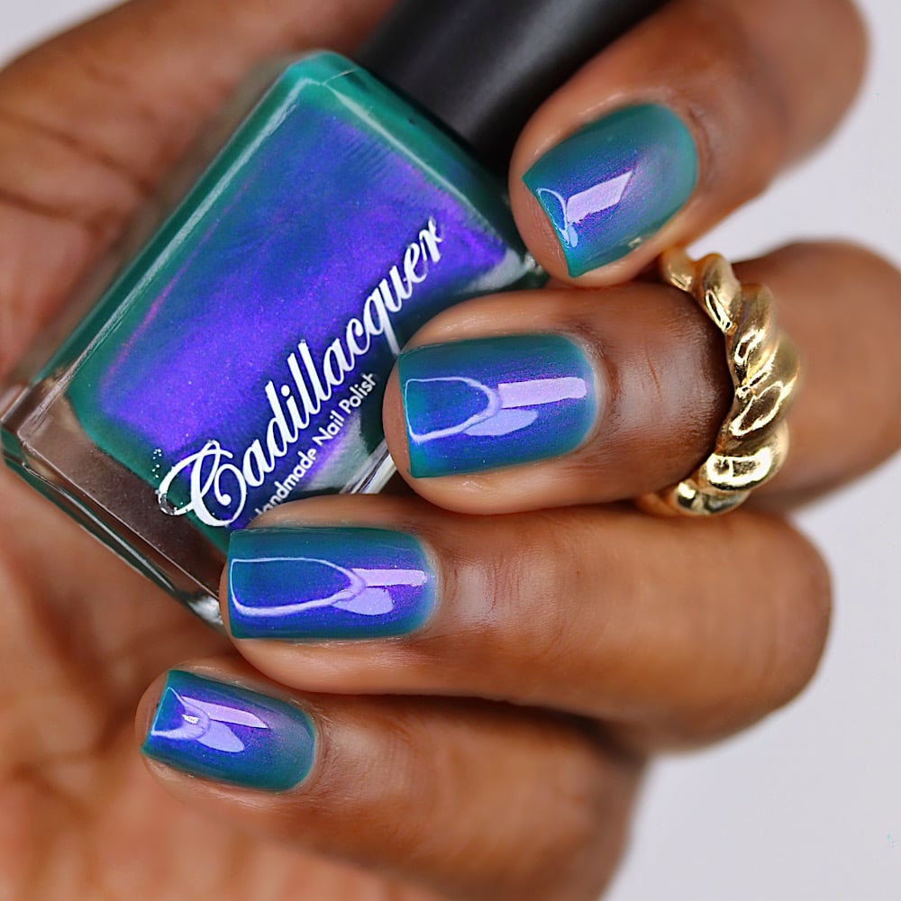 Multichrome shimmer Cadillacquer nail polish in It's All For You, shown in bottle held in hand with matching swatch on four short square nails, shifting from deep teal green base to blue-violet and cyan-teal at angles, on deep skin tone with gold ring accent, studio lighting, swatched by @lacquer_is_life