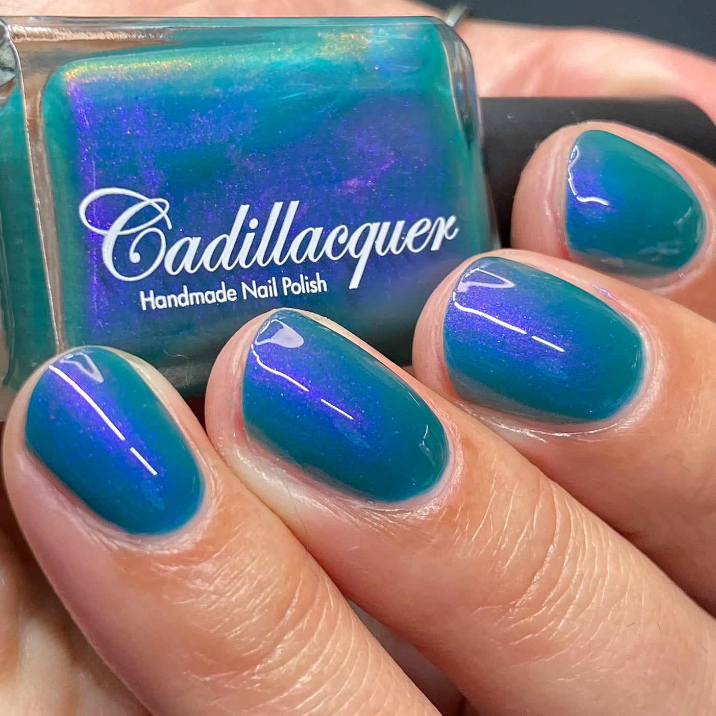 Multichrome shimmer Cadillacquer nail polish in It's All For You, showing a bottle held in hand alongside four swatched oval nails, the teal-green base shifts to violet-blue shimmer at angular light, light-medium skin tone, studio lighting, swatched by @heeymercedes