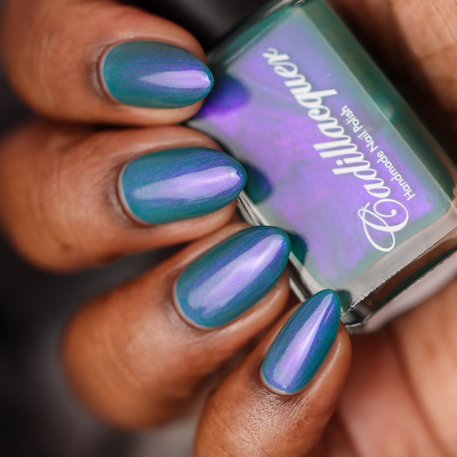 Shimmer multichrome Cadillacquer nail polish in It's All For You, swatched on almond-shaped nails held alongside the bottle, showing a deep teal-green base with a striking purple-to-lavender shimmer shift that sweeps across the nail centre, on deep skin tone, studio lighting, swatched by @doseoflolade