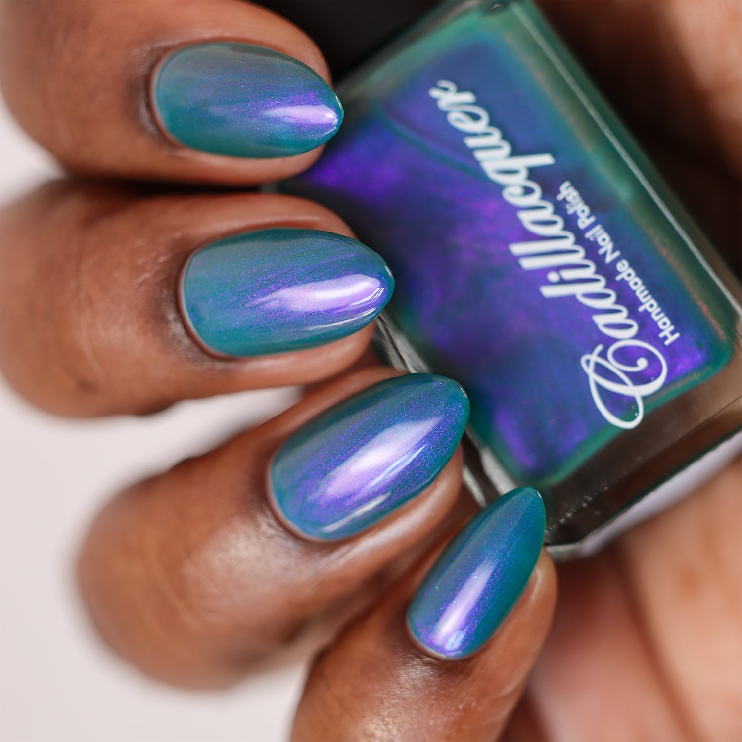 Multichrome shimmer Cadillacquer nail polish in It's All For You, swatched on almond-shaped nails held alongside the bottle, shifting from teal green to violet-purple depending on angle, on deep skin tone, studio lighting, swatched by @doseoflolade