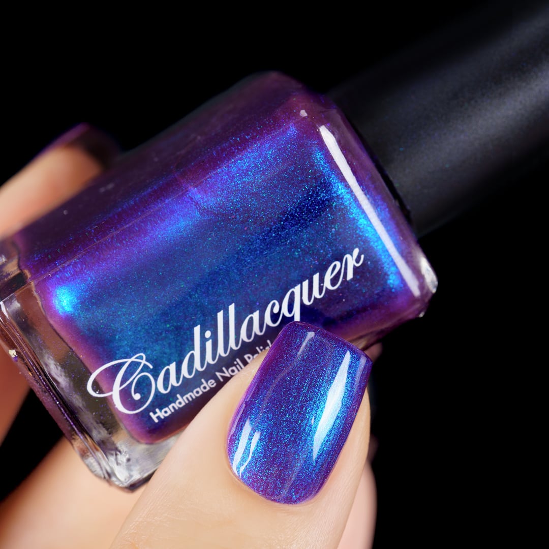 Multichrome shimmer Cadillacquer nail polish in Keep Doubting, shown as a bottle held in hand beside one swatched square nail, the bottle face shifts dramatically from deep violet through electric blue to teal-green shimmer while the nail reads medium purple-violet with blue shimmer flash, light skin tone, studio lighting against black background, swatched by @yyulia_m