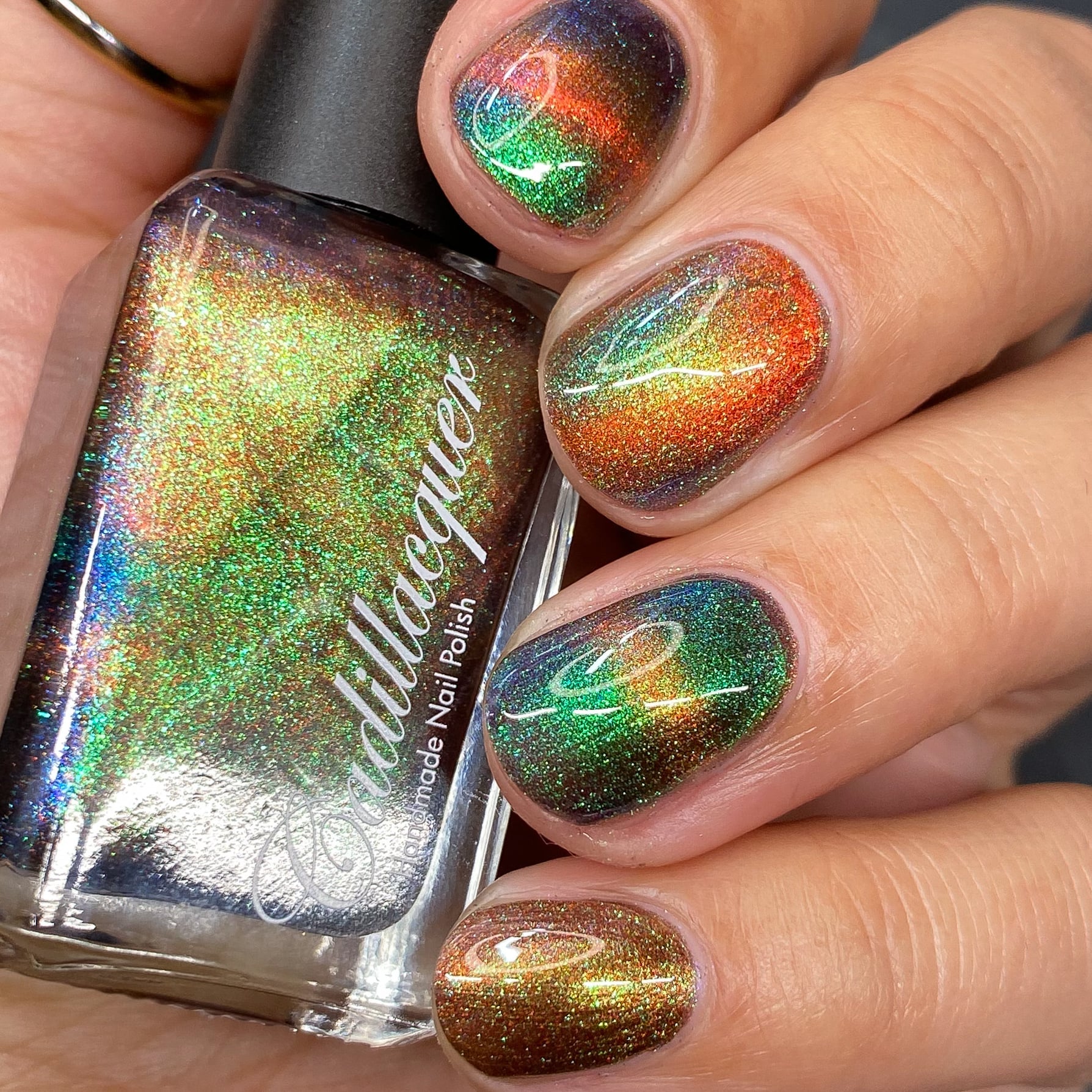 Magnetic multichrome holographic Cadillacquer nail polish in Once You See It, shown on five short natural nails with a swirl magnetic pattern and the matching bottle, the field reads very dark reddish-brown while a vivid copper-orange magnetic band arcs across each nail, with green-to-blue aurora shimmer flashing throughout, light-medium skin tone, studio lighting, swatched by @heeymercedes