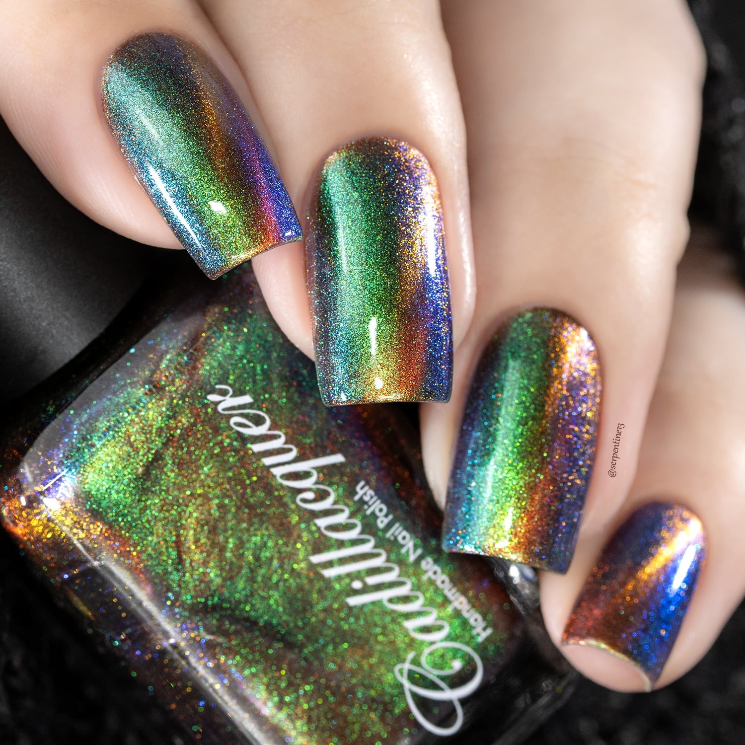 Holographic magnetic multichrome Cadillacquer nail polish in Once You See It, swatched on four square nails with a diagonal amber-gold magnetic band sweeping across deep olive-green and cobalt-blue shifting fields, packed with fine holographic particles that scatter the full spectrum, light skin tone, studio lighting with bottle visible in foreground, swatched by @serpentine13