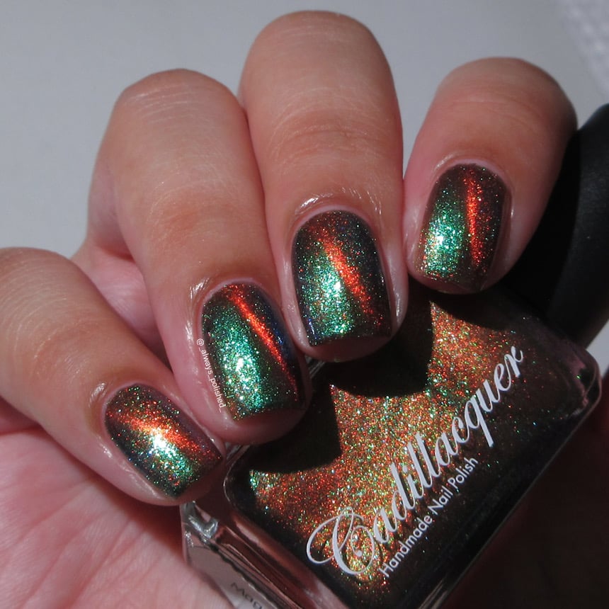 Magnetic multichrome Cadillacquer nail polish in Once You See It, swatched on short natural squoval nails showing a vivid orange magnetic cat-eye stripe over a near-black base with green-to-teal aurora shimmer throughout, light-medium skin tone, indoor warm lighting, swatched by @_always_polished_