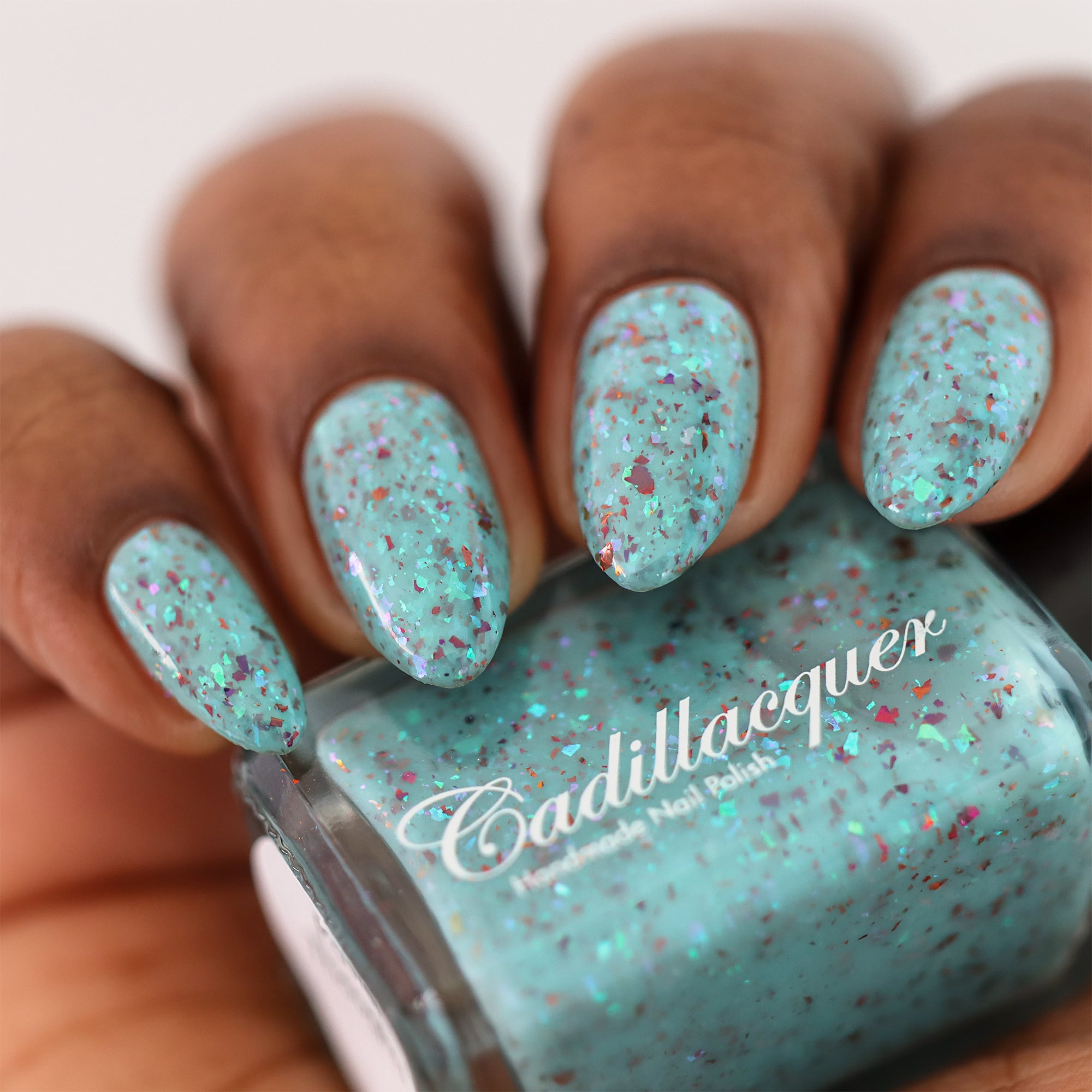 Cadillacquer Pastel Thoughts pastel teal purple chameleon iridescent crelly nail polish, bottle held with four-nail swatch, medium-deep skin, studio lighting