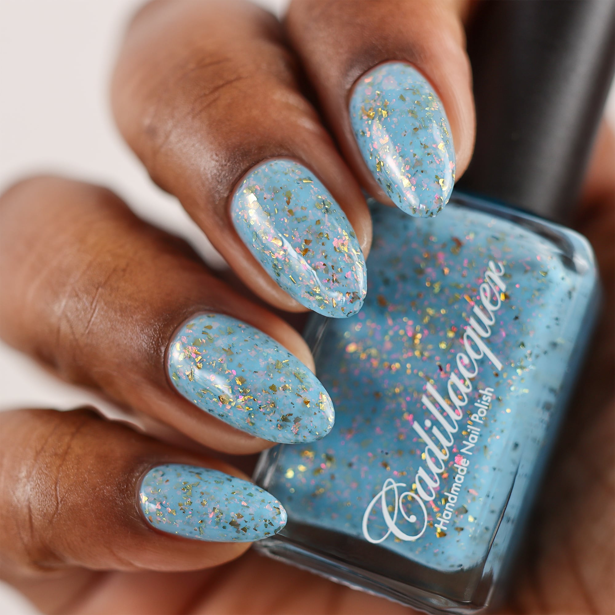 Cadillacquer Slow Mornings light blue gold chameleon iridescent crelly nail polish, bottle held with four-nail swatch, rich skin, studio lighting
