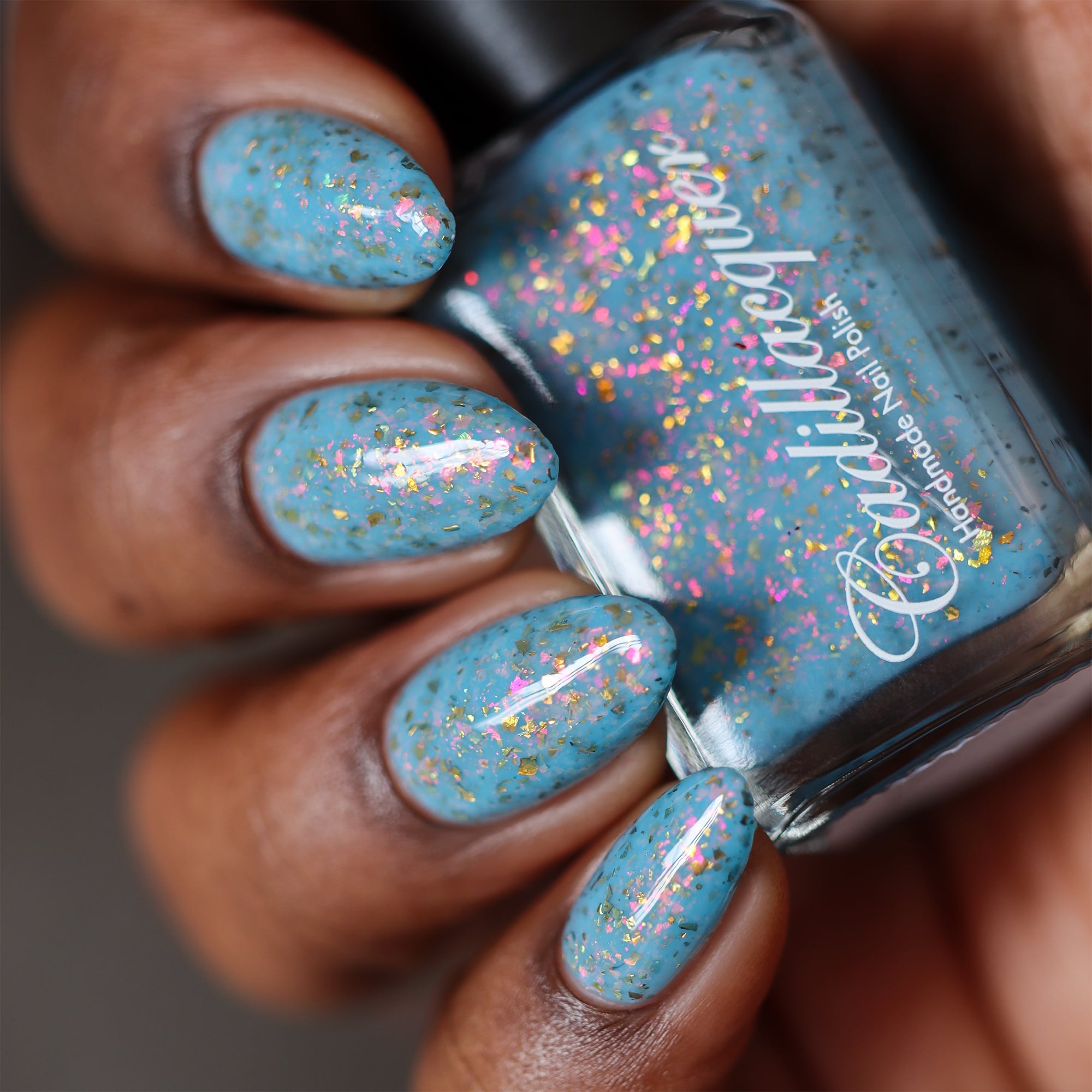 Cadillacquer Slow Mornings light blue gold chameleon iridescent crelly nail polish, bottle held with five-nail swatch, rich skin, studio lighting