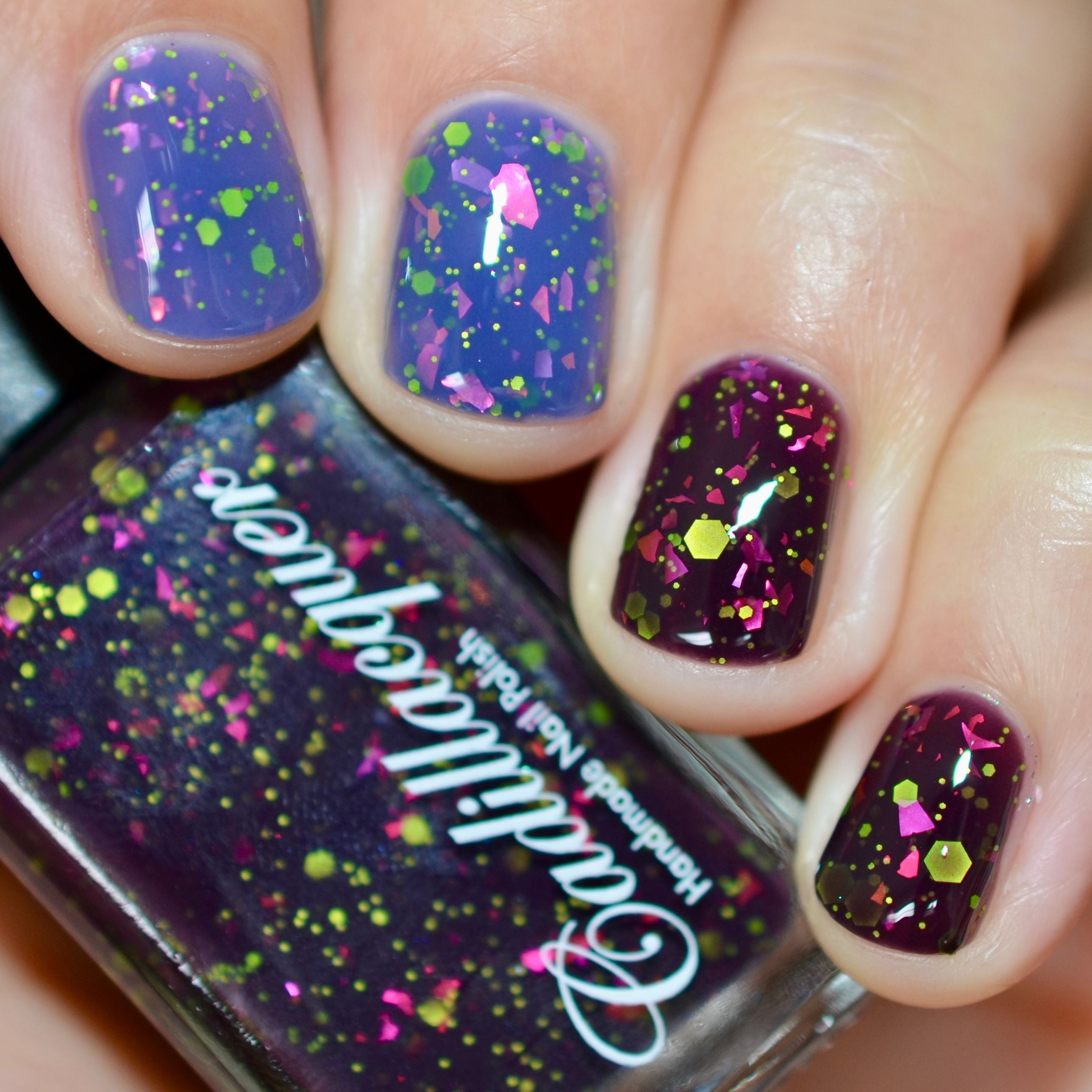 Thermal glitter and flake Cadillacquer nail polish in Strange Happenings, shown as bottle in hand with four swatched nails displaying cold blackened eggplant and warm dusty periwinkle-lavender thermal transition, loaded with neon yellow-green matte glitter in multiple sizes and large hot pink iridescent flakes, on light skin tone, studio lighting, swatched by @caitlinswatches