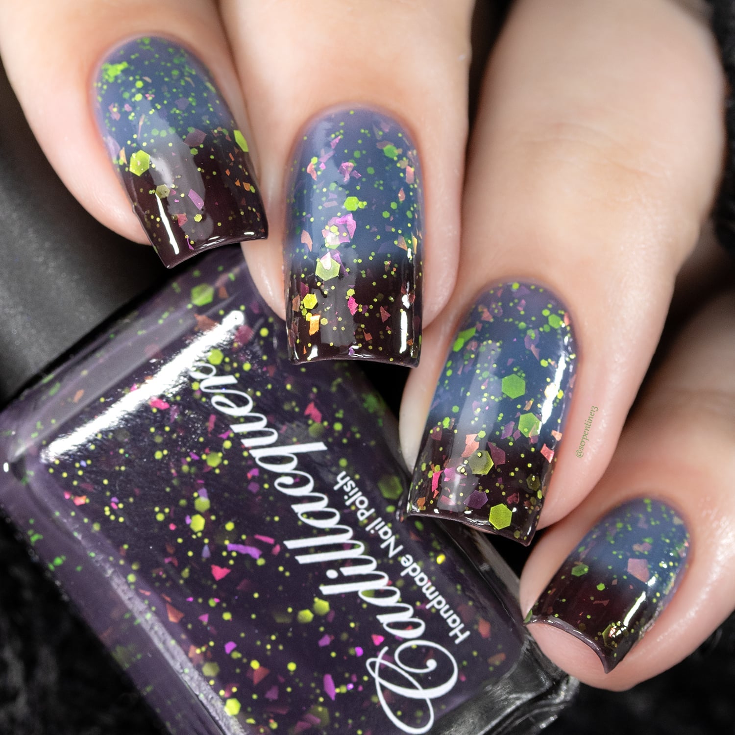 Thermal glitter-and-flake Cadillacquer nail polish in Strange Happenings, shown with bottle in hand and five swatched nails displaying a gradient from blackened eggplant at the cuticle to dusty blue-grey at the tips, loaded with matte neon green glitter in multiple sizes and pink-to-orange iridescent flake pieces, light skin tone, studio lighting, swatched by @serpentine13