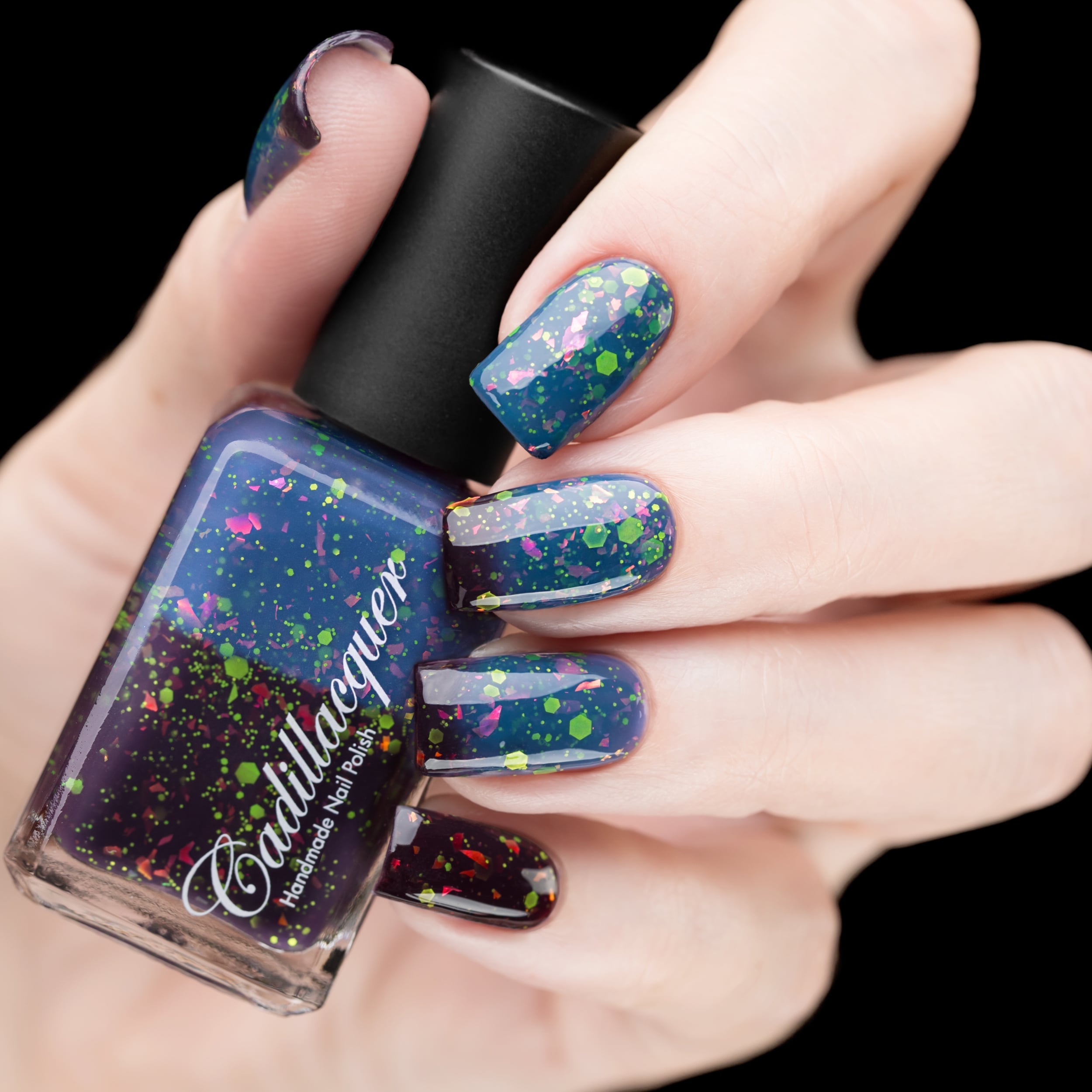 Thermal glitter and flake Cadillacquer nail polish in Strange Happenings, shown as a bottle held in hand alongside four swatched square nails displaying a transition from blackened eggplant purple (cold, at nail tips and bottle) to dusty teal-blue (warm, at nail bases), packed with neon yellow-green matte glitter dots and pink-to-orange iridescent flakes, light skin tone, studio lighting against black background, swatched by @rafinails
