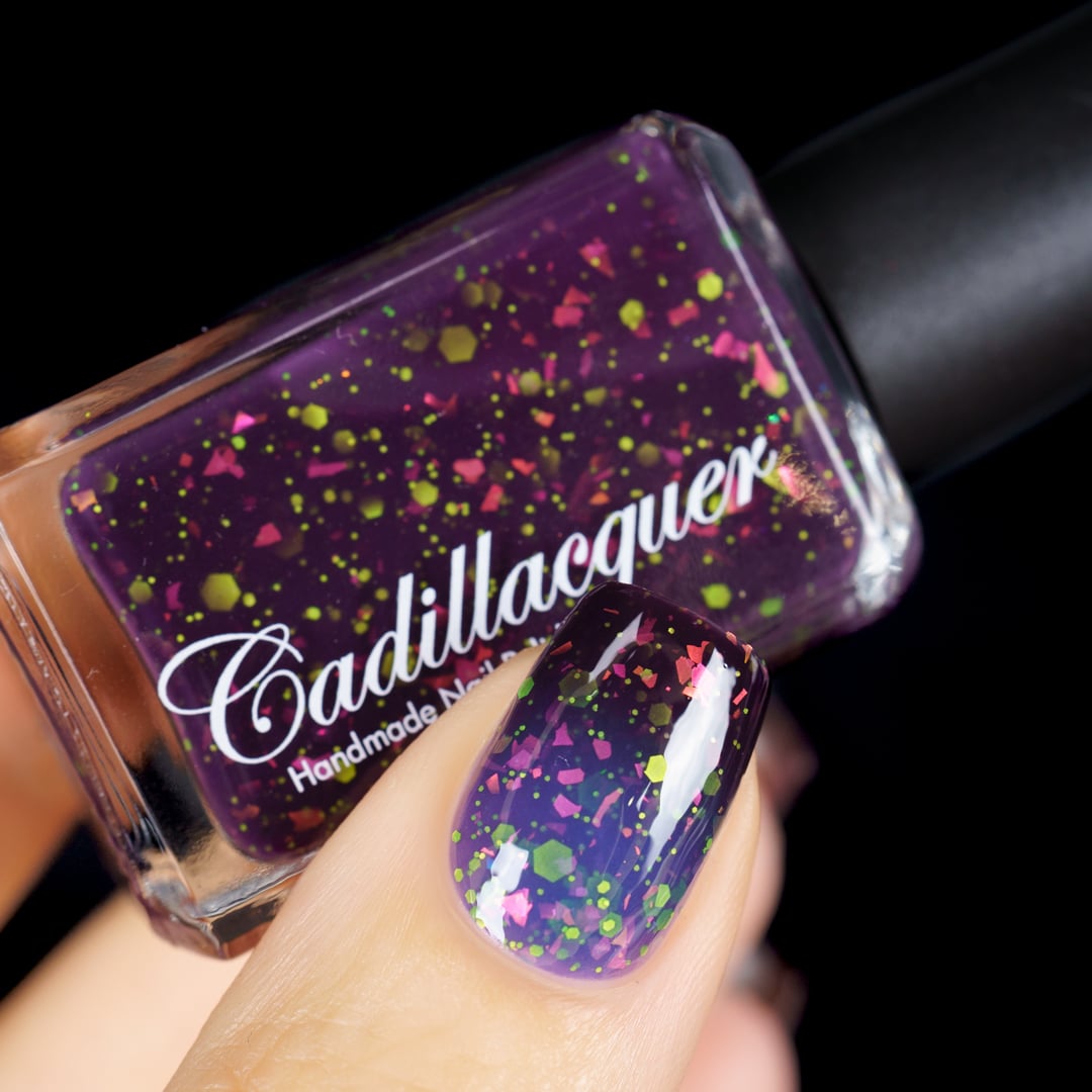 Thermal glitter and iridescent flake Cadillacquer nail polish in Strange Happenings, shown as a bottle-in-hand with one swatched nail on light skin, the blackened eggplant purple base is densely packed with large neon yellow-green matte glitter in varied hex and dot sizes and hot pink iridescent flakes, only the cold thermal state is visible in this frame, studio lighting on black background, swatched by @yyulia_m