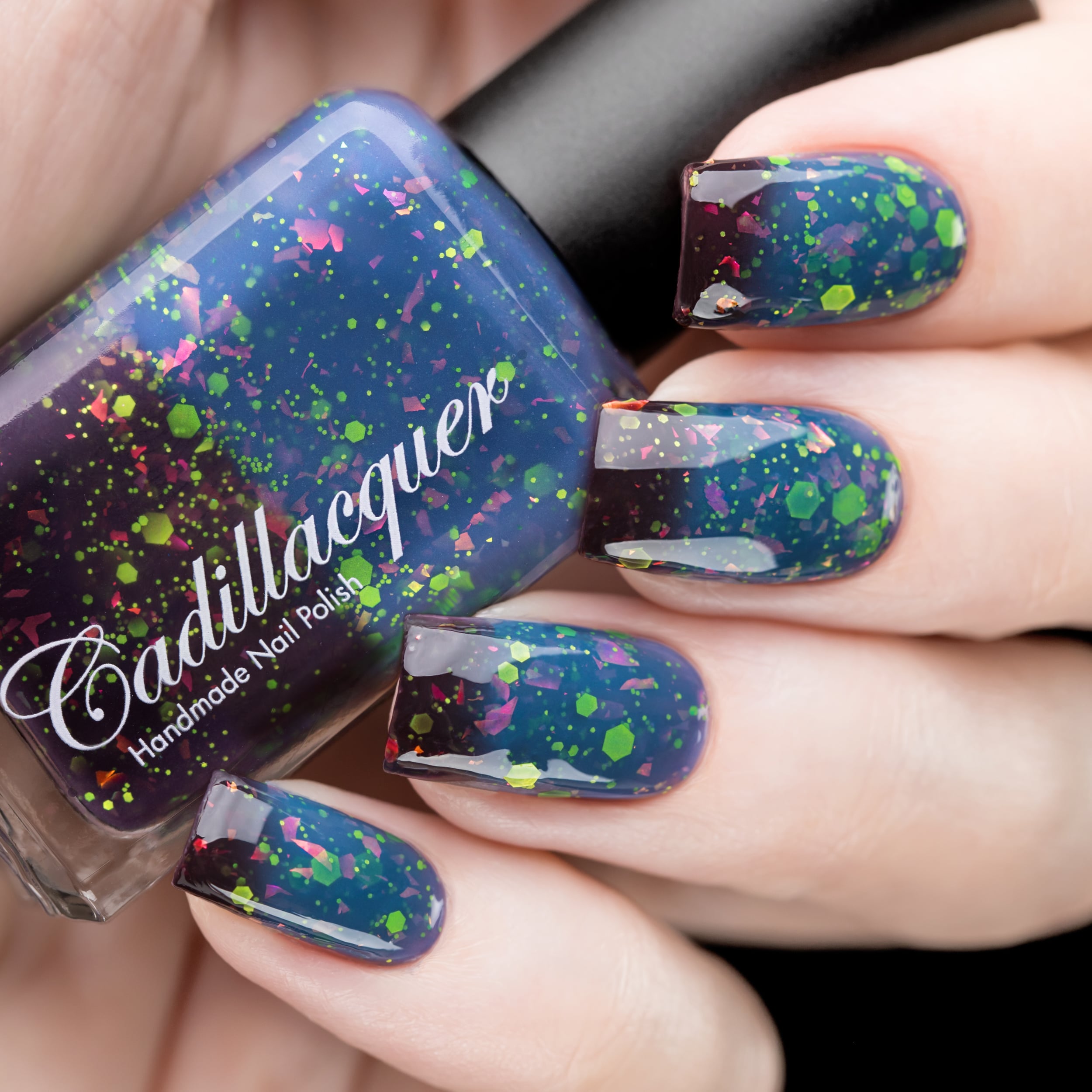 Thermal glitter and iridescent flake Cadillacquer nail polish in Strange Happenings, shown in bottle held alongside five swatched nails transitioning from blackened eggplant cold state to dusty teal-blue warm state, loaded with matte neon green hex and dot glitter and pink-to-orange iridescent flakes, light skin tone, studio lighting, swatched by @rafinails