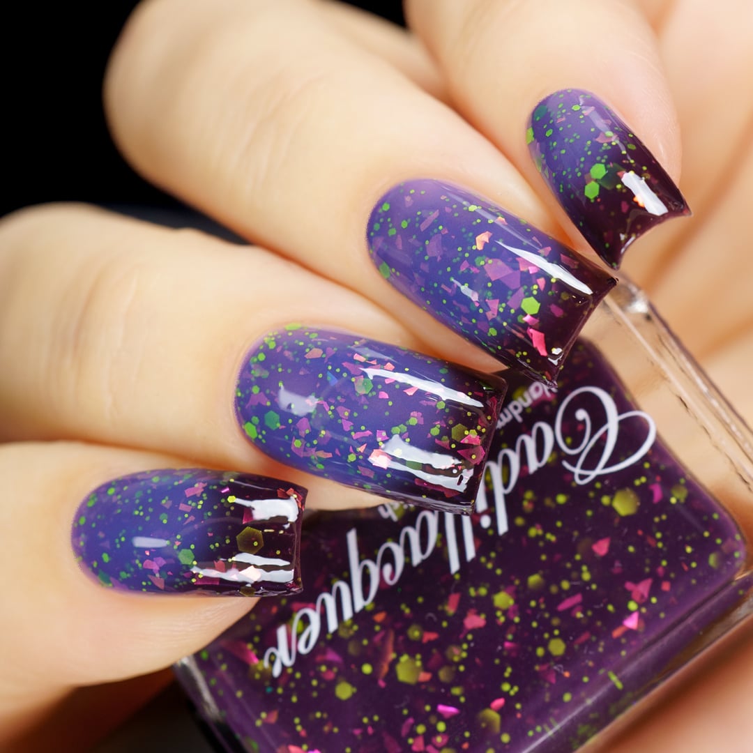 Thermal glitter and flake Cadillacquer nail polish in Strange Happenings, shown in a bottle-in-hand swatch with four almond-shaped nails transitioning from blackened eggplant purple at the tips (cold) to dusty blue-violet at the base (warm), loaded with hot pink iridescent flakes and matte neon green glitter in multiple sizes, light skin tone, studio lighting, swatched by @yyulia_m
