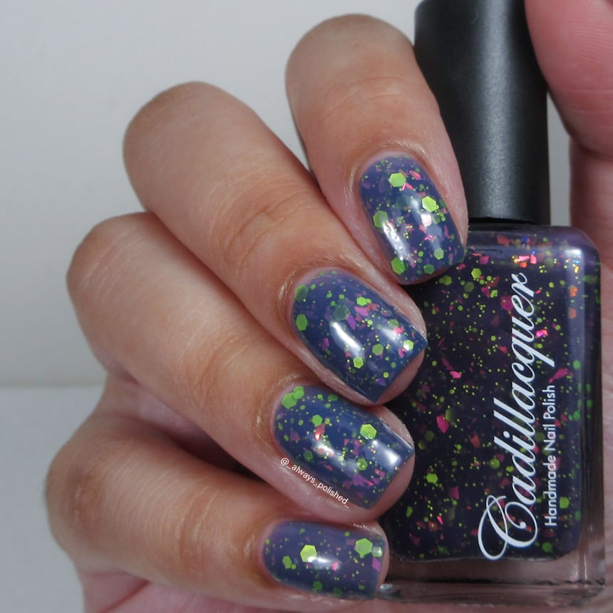 Thermal glitter-and-flake Cadillacquer nail polish in Strange Happenings, shown swatched on four short natural nails held alongside the bottle, nails display the warm dusty slate-blue state loaded with matte neon green hex and dot glitter and hot-pink iridescent flakes, while the bottle shows the dark blackened eggplant cold state, light skin tone, studio lighting, swatched by @_always_polished_