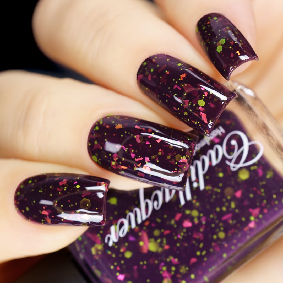 Thermal glitter-and-flake Cadillacquer nail polish in Strange Happenings, shown swatched on four medium-length square nails in the cold state, a deep blackened eggplant base densely scattered with matte neon chartreuse green glitter in mixed sizes and hot pink to orange iridescent flakes, a matching bottle is partially visible, on light skin, in studio lighting, swatched by @yyulia_m
