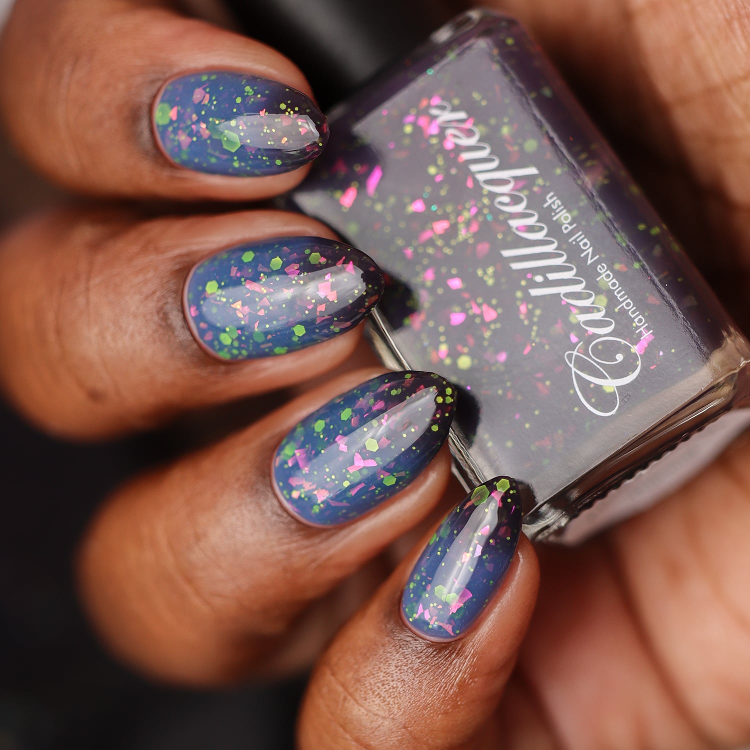 Thermal glitter-and-flake Cadillacquer nail polish in Strange Happenings, swatched on eight oval almond-shaped nails on deep skin tone, showing a dusty blue warm state transitioning to blackened eggplant cold state at cuticles, loaded with large hot-pink iridescent flakes and matte neon green glitter hexagons, photographed in studio lighting with the Cadillacquer bottle held alongside, swatched by @doseoflolade
