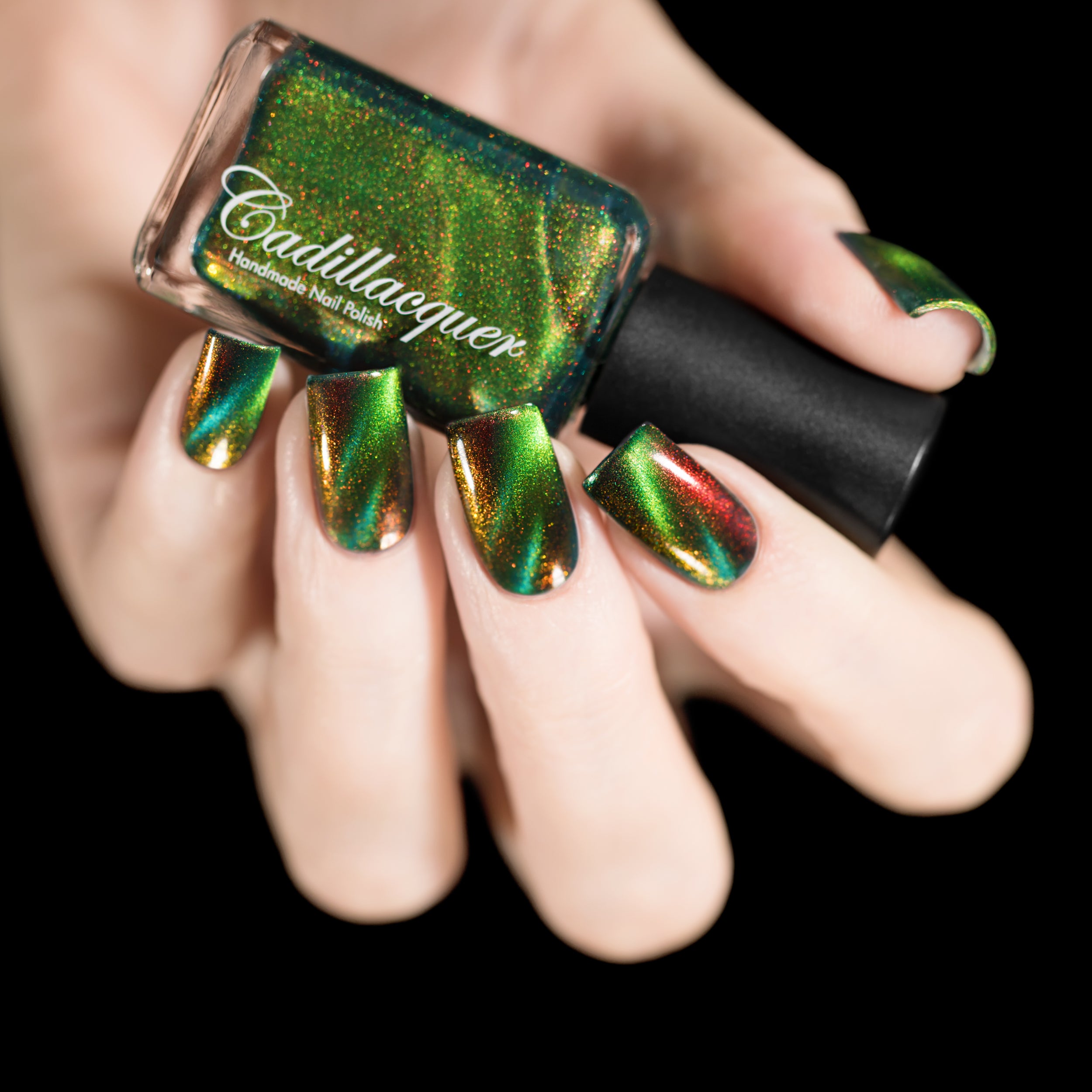 Magnetic multichrome shimmer Cadillacquer nail polish in I Found A Place, a hand holding the bottle over five swatched squoval nails, the nails shifting from dark teal-green in the field through lime green to deep amber-gold along the magnetic band, with red and orange aurora shimmer flashing at angles, light skin tone, studio lighting, swatched by @rafinails