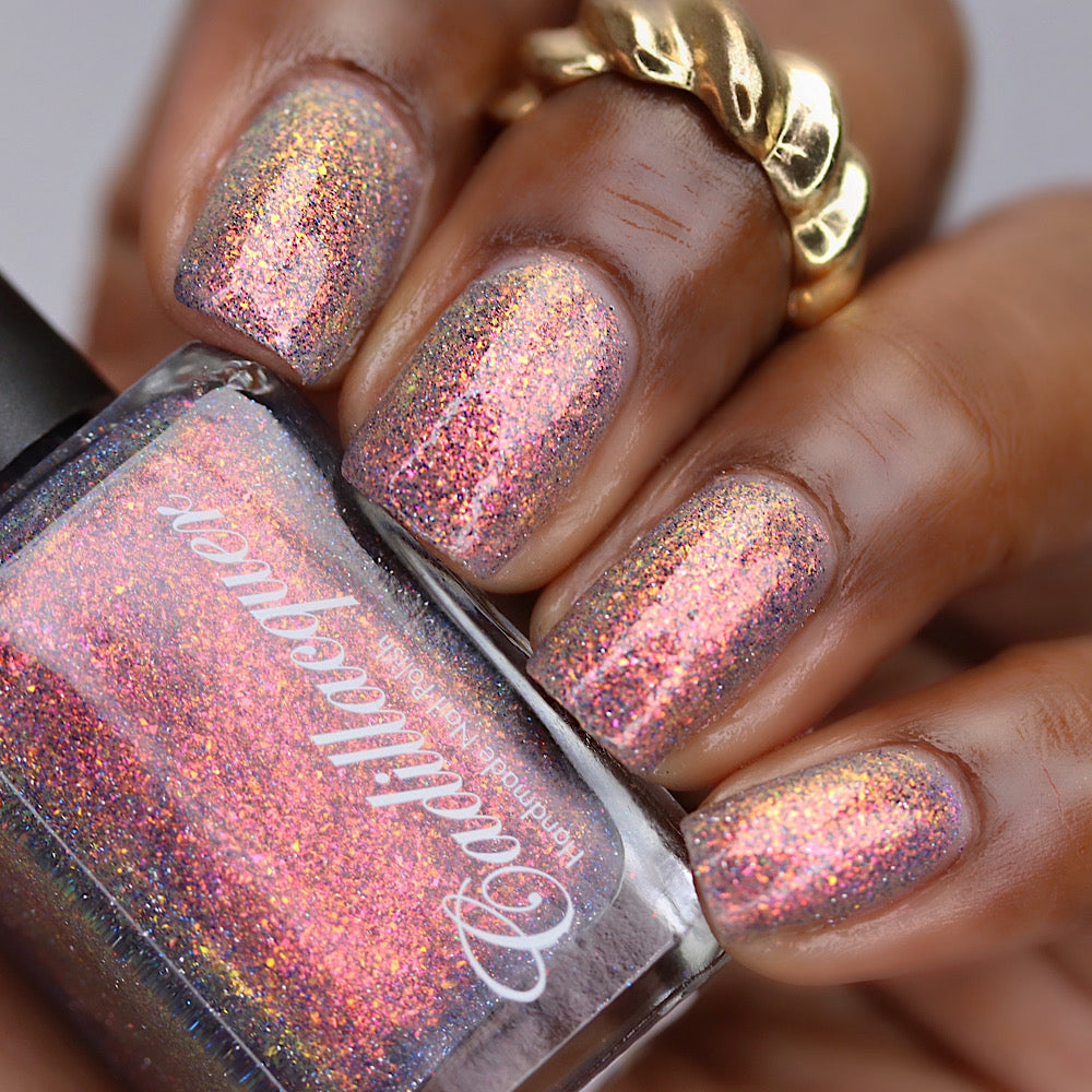 Iridescent multichrome micro-flake and reflective glitter Cadillacquer nail polish in You Wished For This, shown swatched on five short natural nails with a sheer mauve-grey base packed with warm orange-to-pinkish-red-to-golden-yellow iridescent micro flakes and scattered charcoal and blue holographic micro-glitter, on deep skin tone with a gold ring accent, studio lighting, swatched by @lacquer_is_life