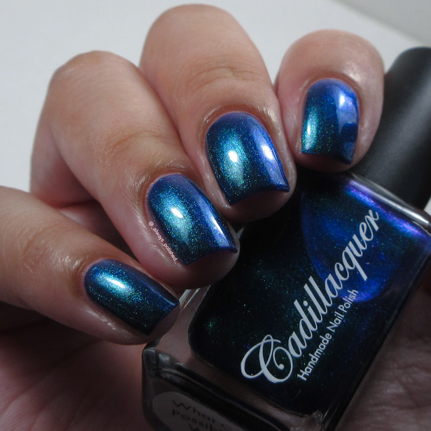 Multichrome shimmer Cadillacquer nail polish in What Could Possibly Go Wrong?, swatched on eight short natural nails held alongside the dark bottle, shifting from deep indigo base through teal-green to cobalt-blue depending on angle, on light-medium skin, indoor warm lighting, swatched by @_always_polished_