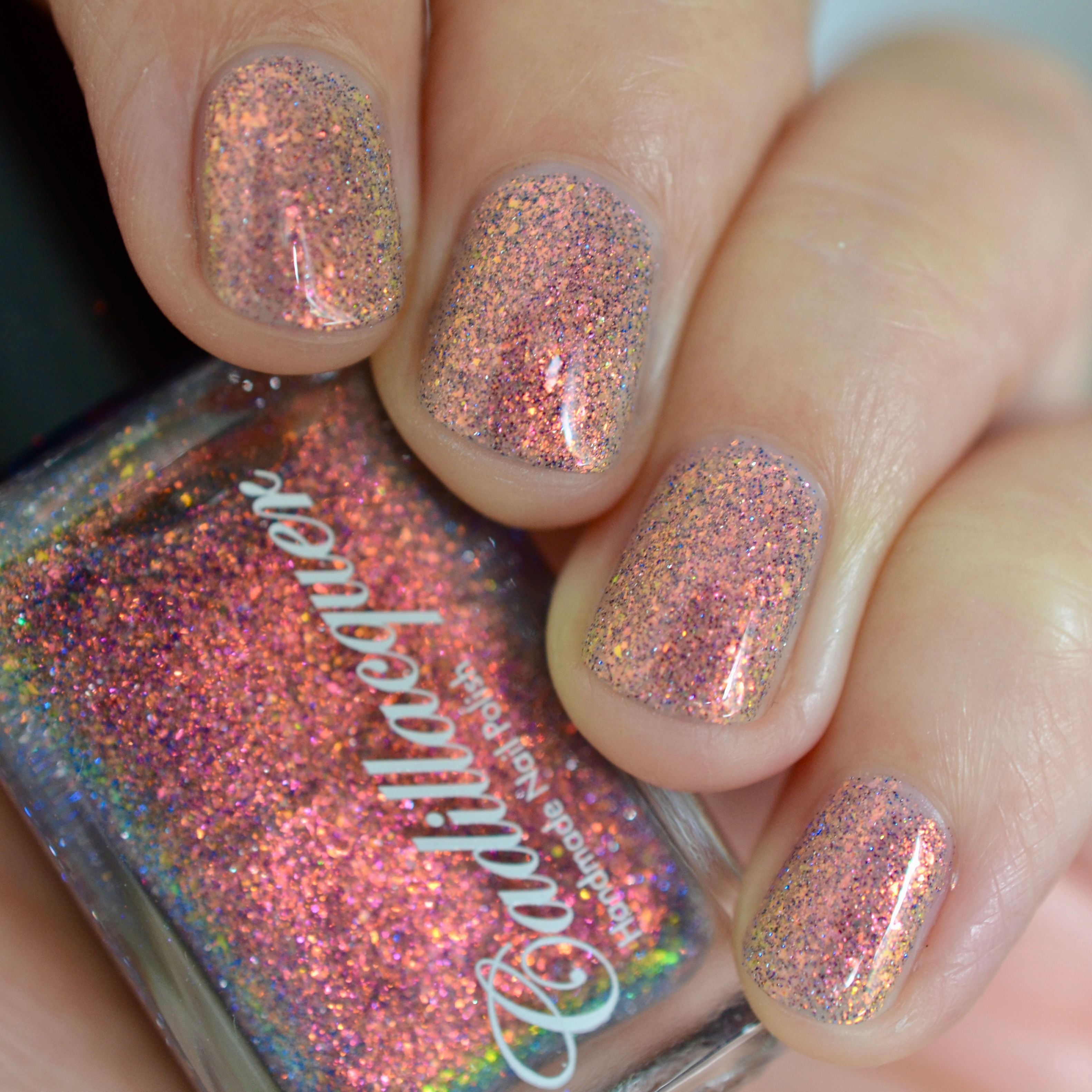 Iridescent micro-flake and holographic glitter Cadillacquer nail polish in You Wished For This, shown as a swatch on five short squoval nails alongside the polish bottle, flakes shift from warm coral-orange through red-pink to golden yellow with scattered blue-violet and charcoal holographic micro glitter in a sheer base, light-medium skin tone, studio lighting, swatched by @caitlinswatches
