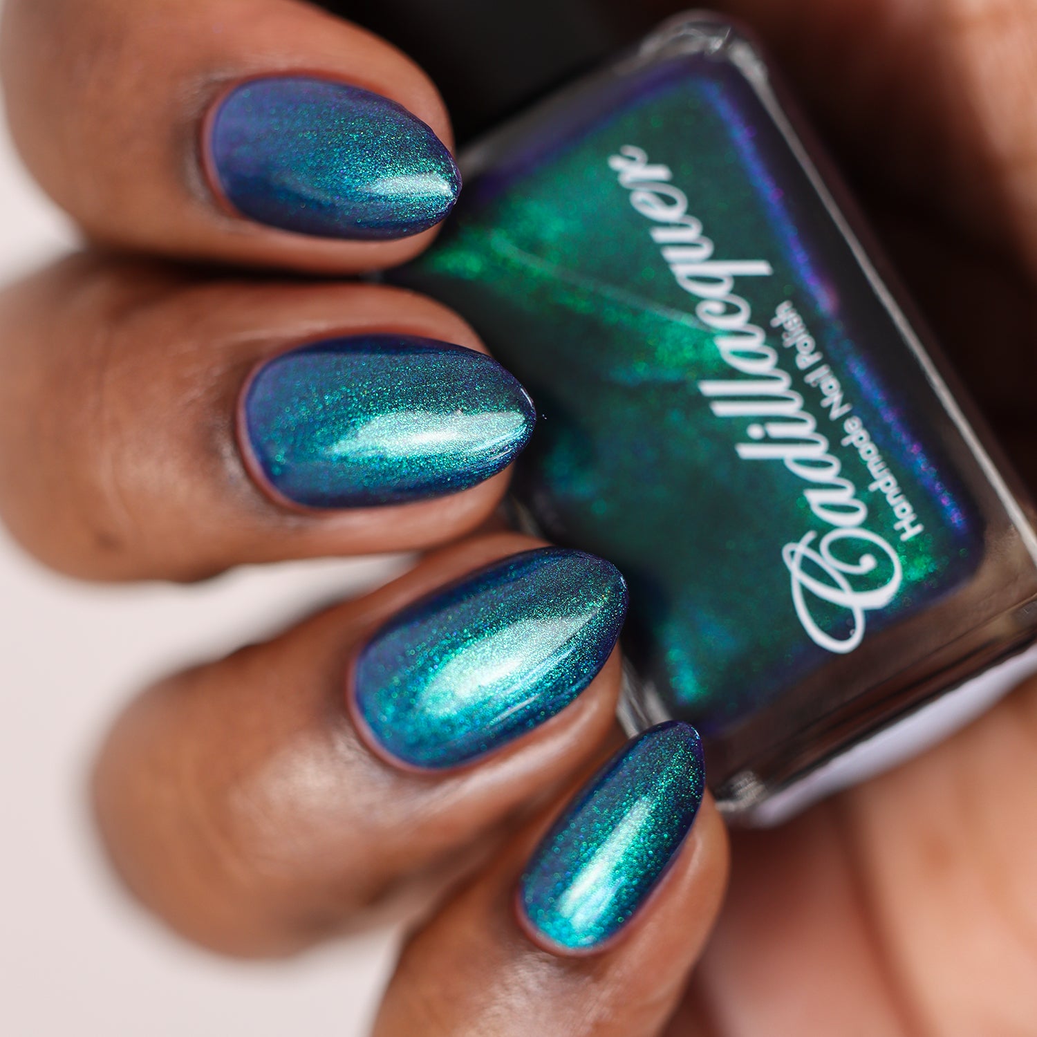 Multichrome shimmer Cadillacquer nail polish in What Could Possibly Go Wrong?, shown swatched on five almond-shaped nails holding a Cadillacquer bottle, shifting from deep indigo base through cobalt blue to vivid teal-green depending on viewing angle, on deep skin tone, studio lighting, swatched by @doseoflolade