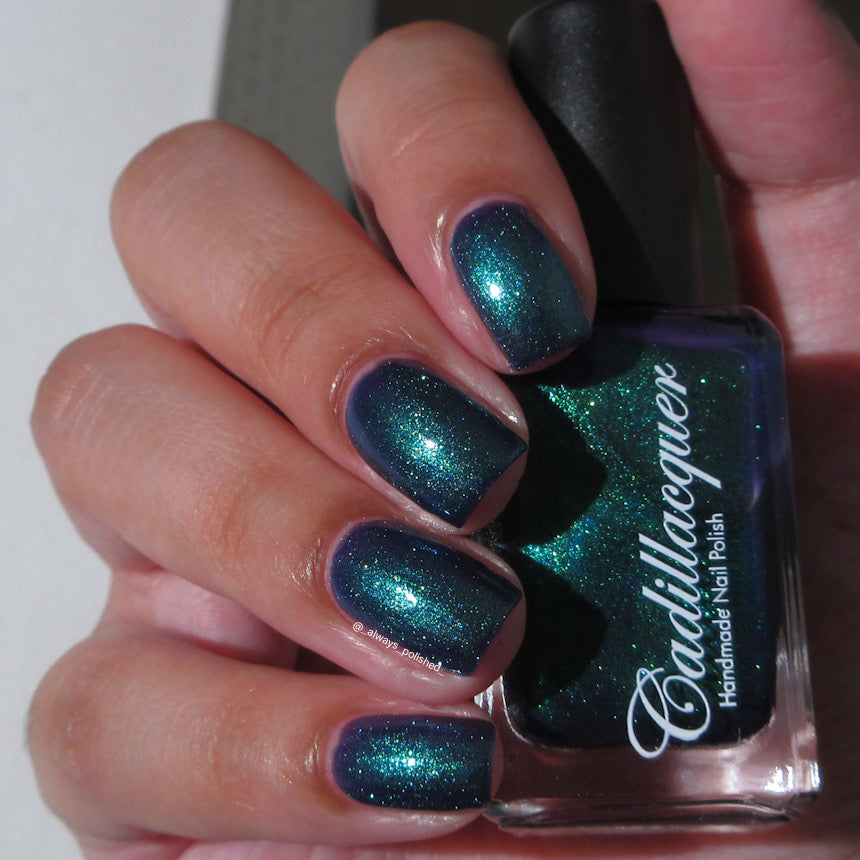 Multichrome shimmer Cadillacquer nail polish in What Could Possibly Go Wrong?, swatched on eight short natural nails held alongside the bottle, shifting from deep indigo base through vivid teal-green shimmer at angle, light-medium skin tone, indoor warm lighting, swatched by @_always_polished_