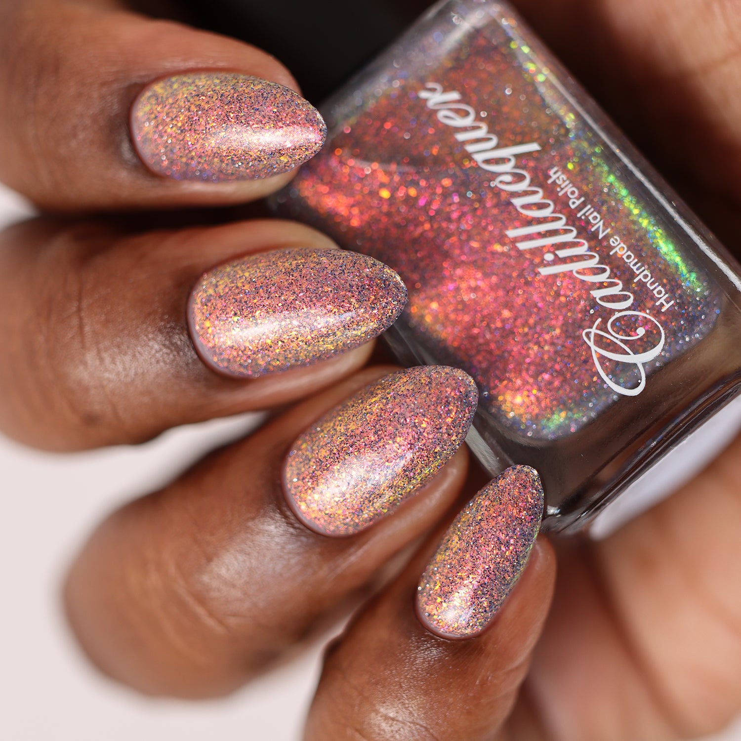 Holographic iridescent micro-flake Cadillacquer nail polish in You Wished For This, shown swatched on five almond-shaped nails on a rich skin tone hand holding the bottle, with warm peachy rose-copper base packed with red-orange to golden-yellow micro flakes and scattered charcoal and blue holographic glitter particles, studio lighting, swatched by @doseoflolade