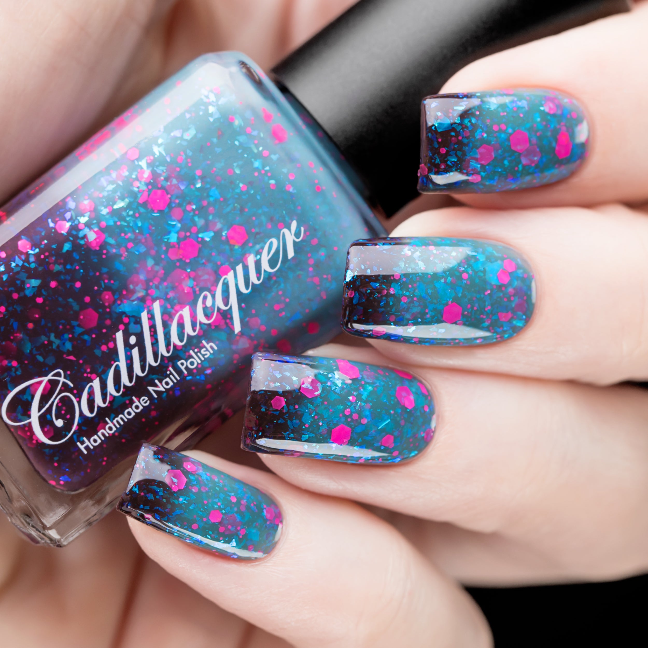 Thermal glitter-and-flake Cadillacquer nail polish in What Lurks Within, shown as bottle in hand with four swatched nails displaying deep blackened eggplant cold zones transitioning to dark teal-blue warm zones, loaded with neon hot pink matte hexagon glitter in multiple sizes and iridescent cyan-blue flakes, light skin tone, studio lighting, swatched by @rafinails