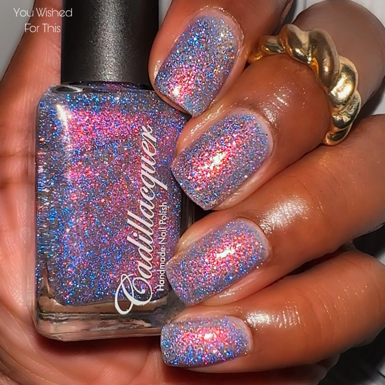 Flake and reflective glitter Cadillacquer nail polish in You Wished For This, shown in bottle held in hand with swatch on five square nails, sheer lavender-grey base packed with pink-to-orange iridescent micro flakes and electric blue reflective glitter that bursts with hot pink-red flash in direct light, medium-deep skin tone, direct flash lighting, swatched by @lacquer_is_life