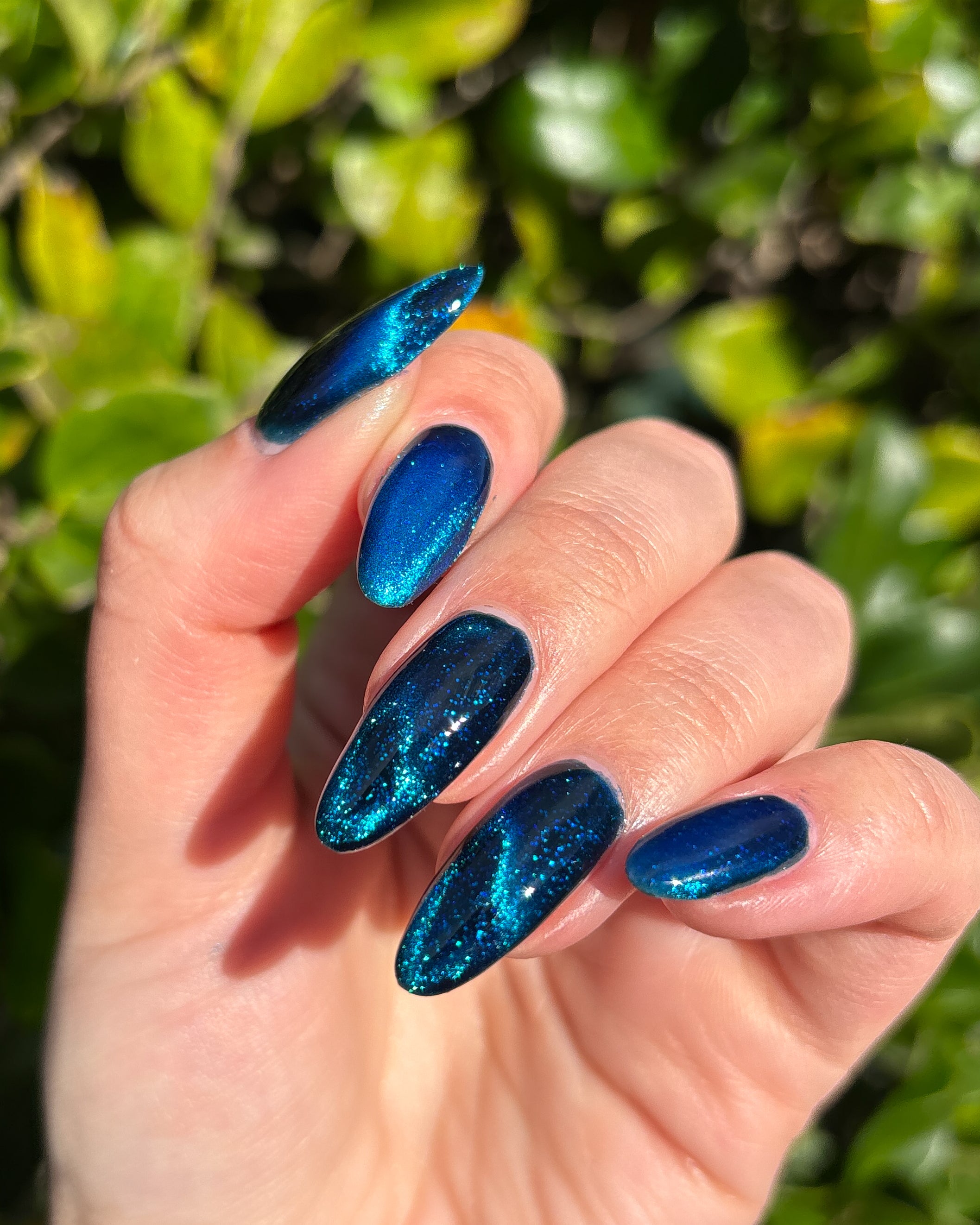 Heart of the Ocean by Glitch Lacquer on five almond nails outdoors against green foliage, deep blue magnetic jelly with blue glitters, light skin tone, bright sunlight.