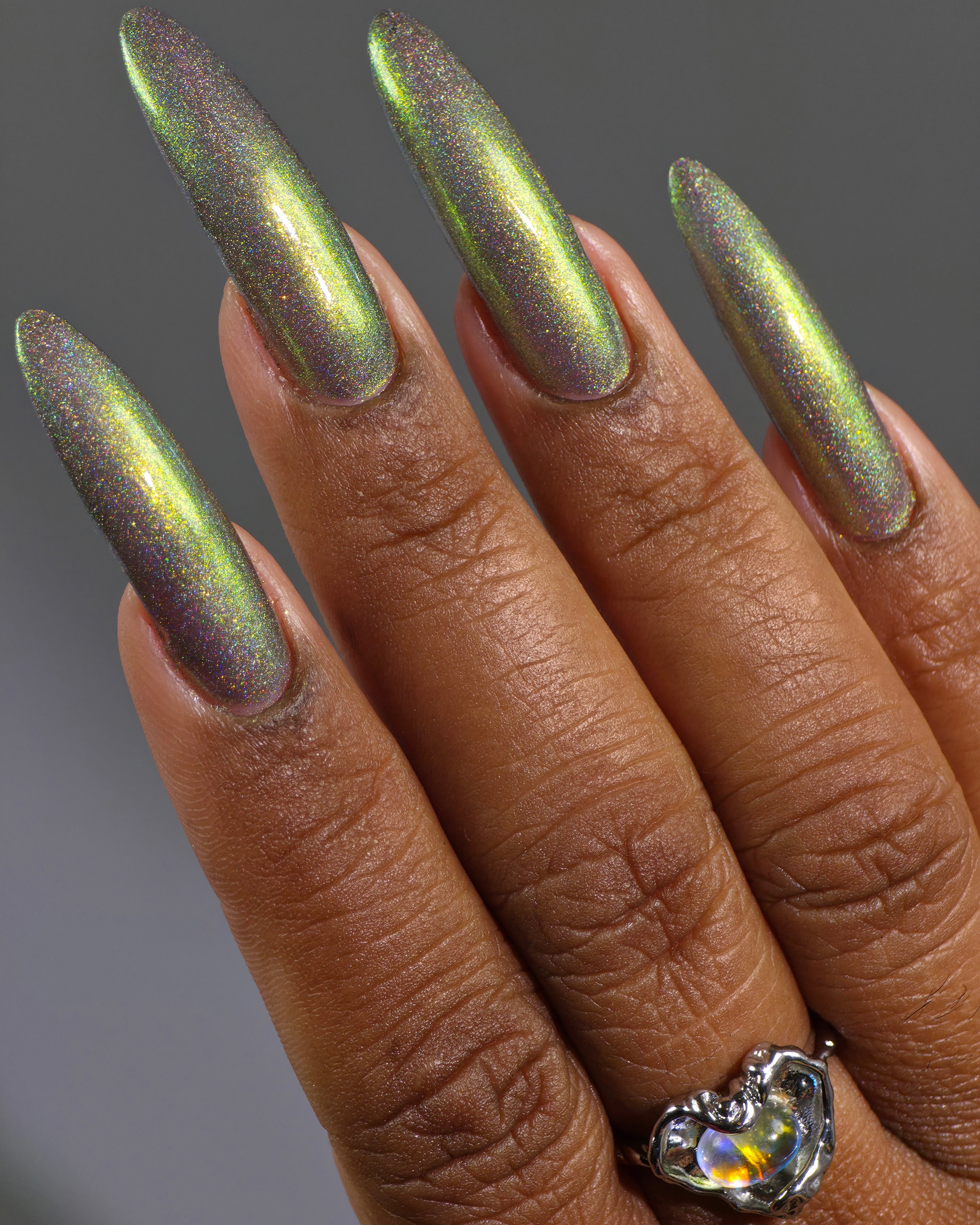 Safe House Glow by Glitch Lacquer, grey base, gold-to-teal multichrome shift, holo shimmer, on deep skin tone stiletto nails
