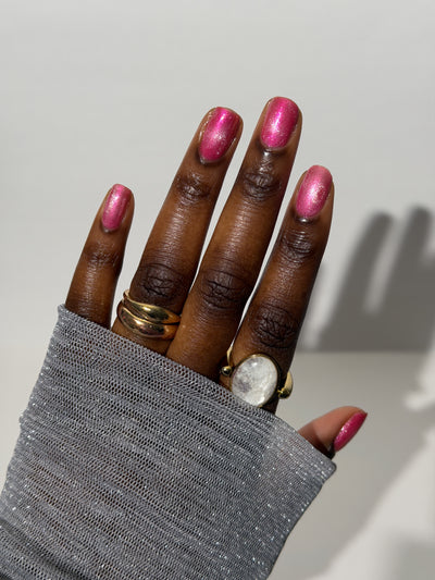 Lovey Dovey by Glitch Lacquer on five round nails, warm pink polish with pink-to-gold reflective shimmer and silver magnetic pigment, deep skin tone, bright studio lighting.