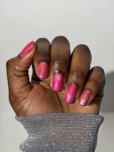 Lovey Dovey by Glitch Lacquer on five round nails showing a subtle magnetic stripe, warm pink polish with pink-to-gold reflective shimmer, deep skin tone, studio lighting.