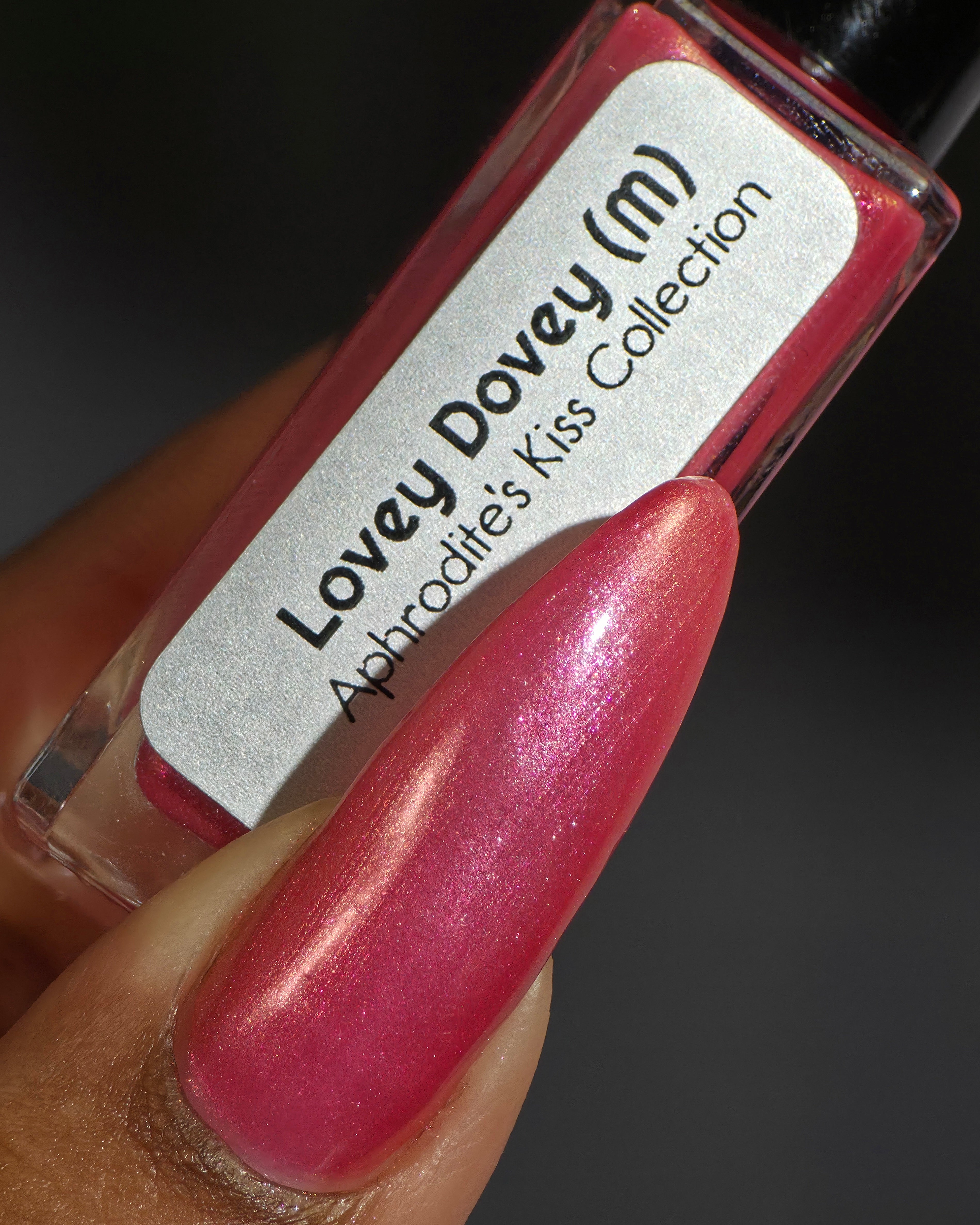Lovey Dovey by Glitch Lacquer bottle held in hand with one long almond nail swatched, warm pink polish with pink-to-gold reflective shimmer and silver magnetic pigment, deep skin tone, studio lighting.
