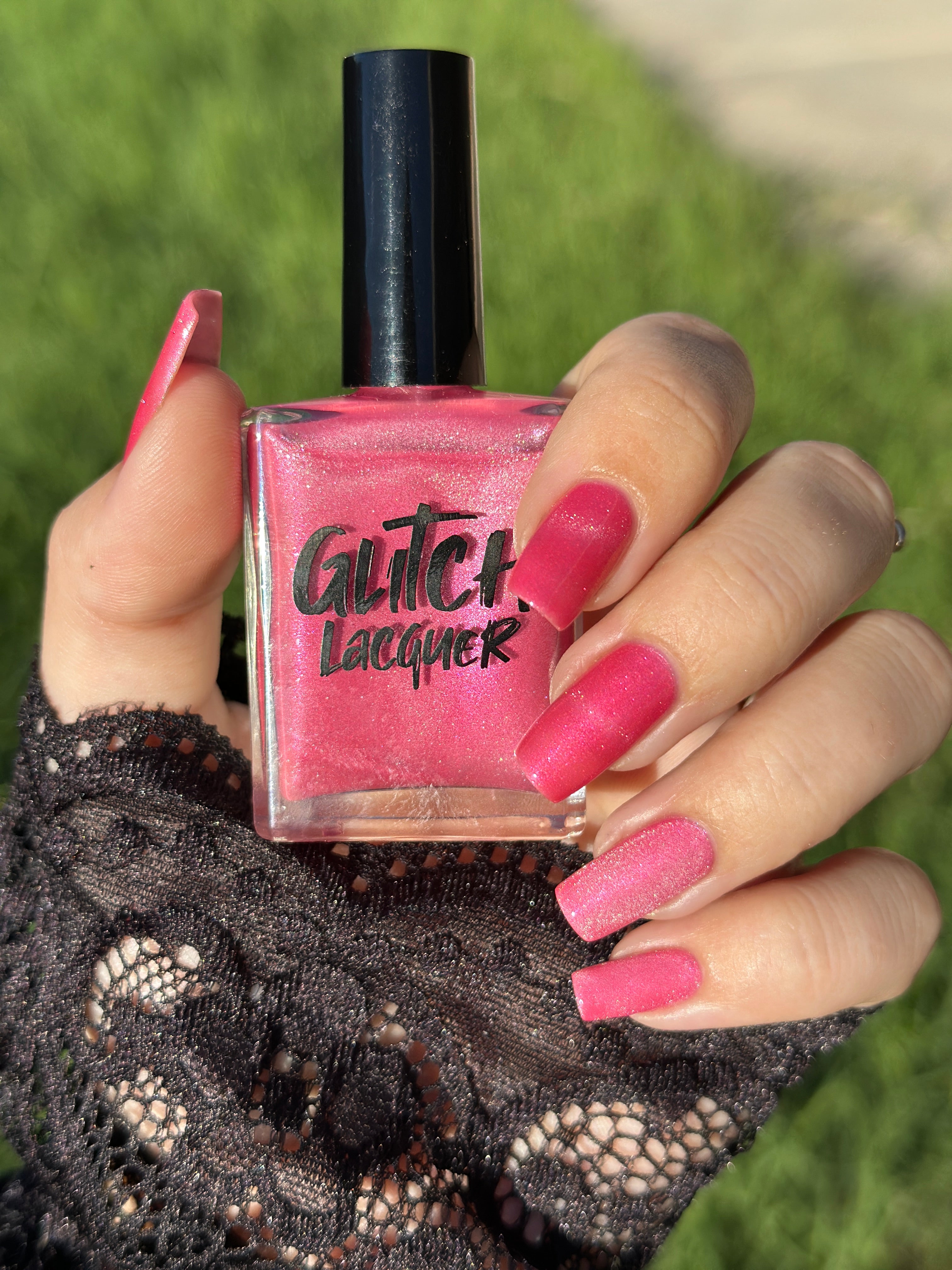 Lovey Dovey by Glitch Lacquer bottle held in hand with five squoval nails swatched showing magnetic stripes, warm pink polish with pink-to-gold reflective shimmer, light-medium skin tone, bright daylight.