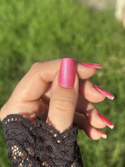 Lovey Dovey by Glitch Lacquer on five rectangular nails worn with a black lace cuff, warm pink polish with pink-to-gold reflective shimmer, light-medium skin tone, bright daylight.